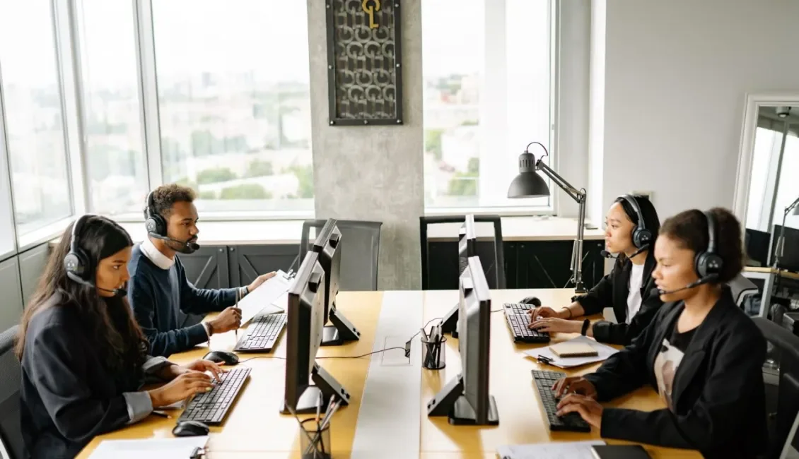 Four people in a modern office working at desks with computers, wearing headsets, engaged in a video conference or customer service call.
