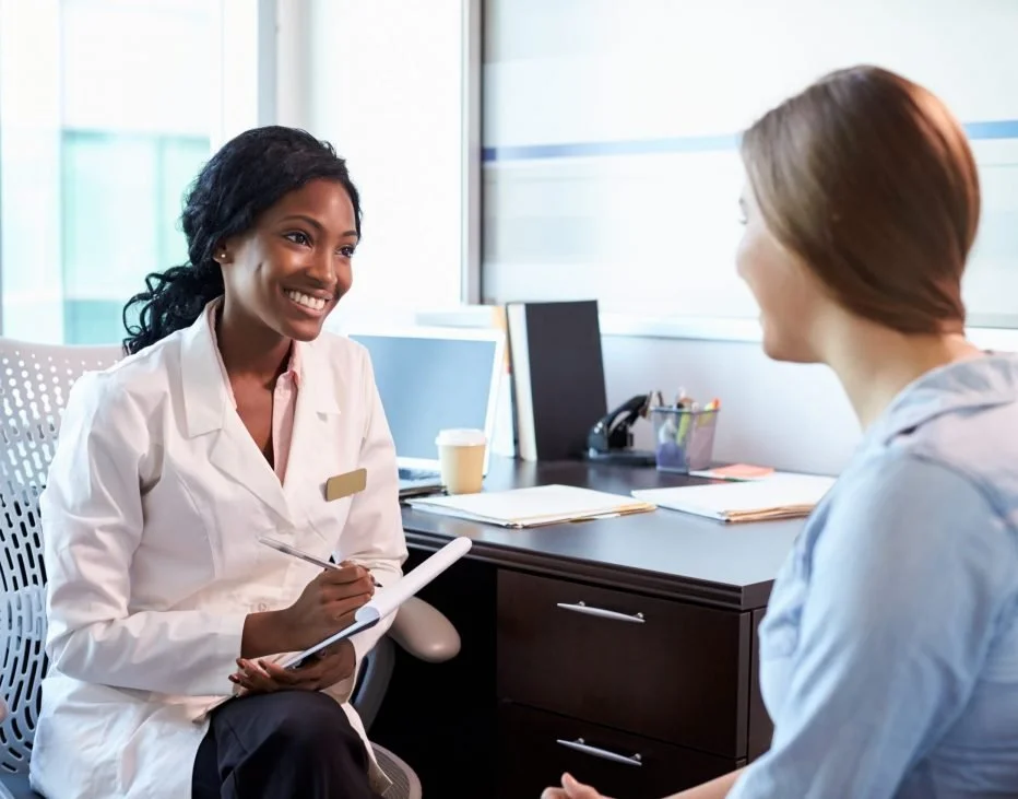 A woman in a white coat sitting across from a patient, smiling and taking notes, in a clinic or medical office.