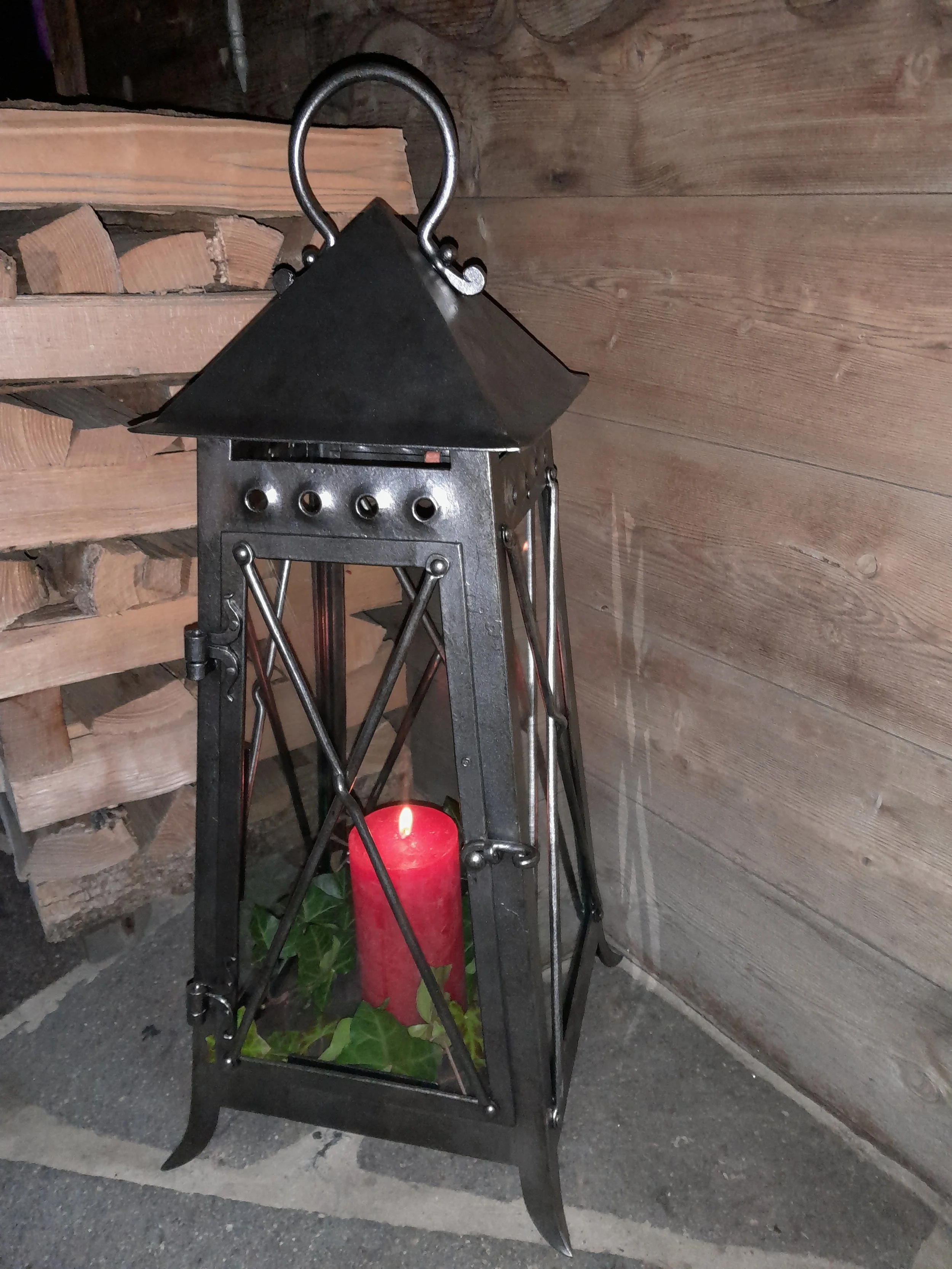 Eine schwarze Laterne mit rotem Kerzenlicht und grünen Blättern im Inneren, steht auf einem Steinboden in einer Holzumgebung.