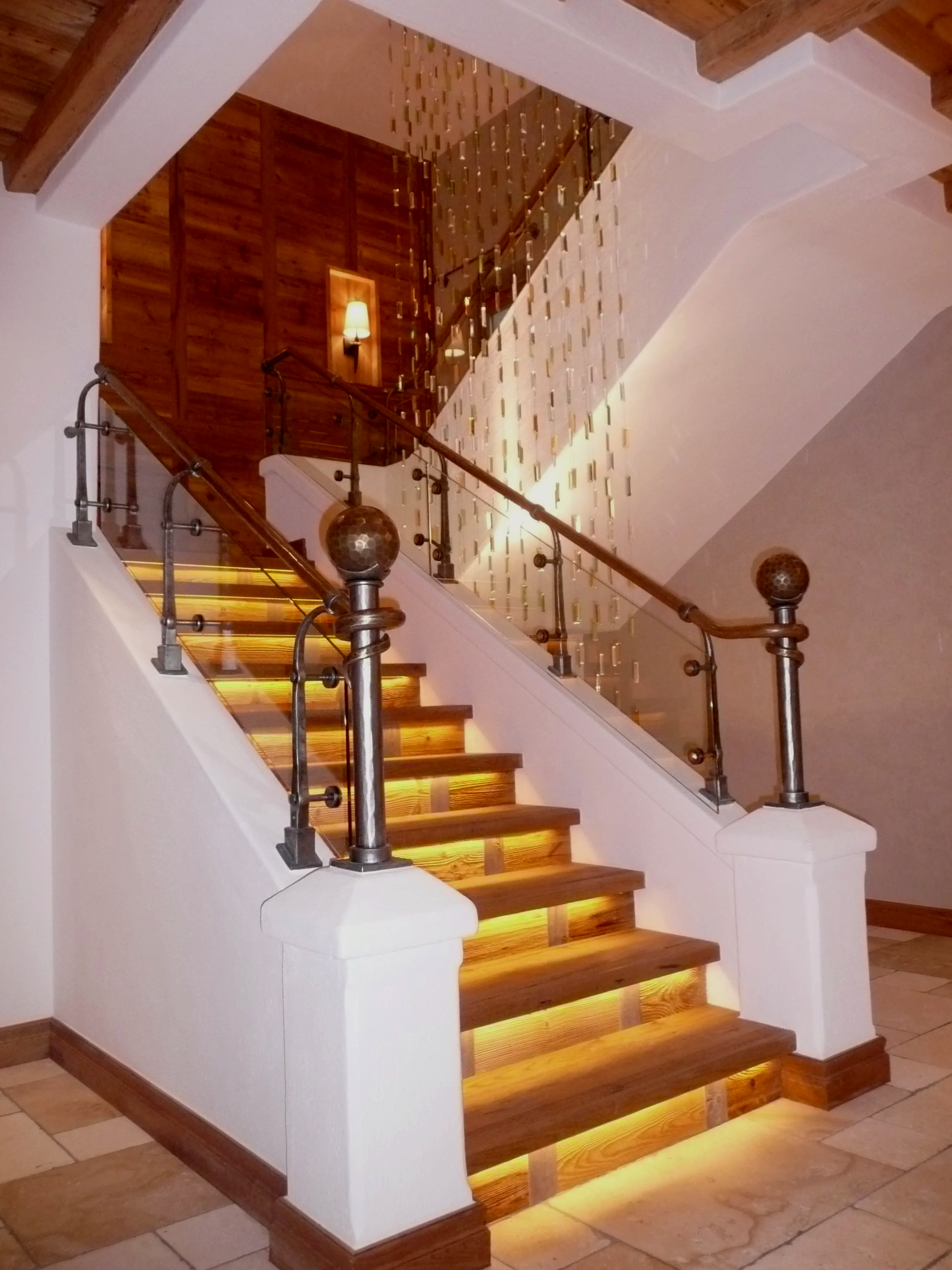 Innenaufnahme einer Holztreppe mit beleuchteten Stufen und einem Geländer aus Metall und Glas, im Hintergrund eine Wand mit Holzverkleidung und einem Wandlampenleuchte.