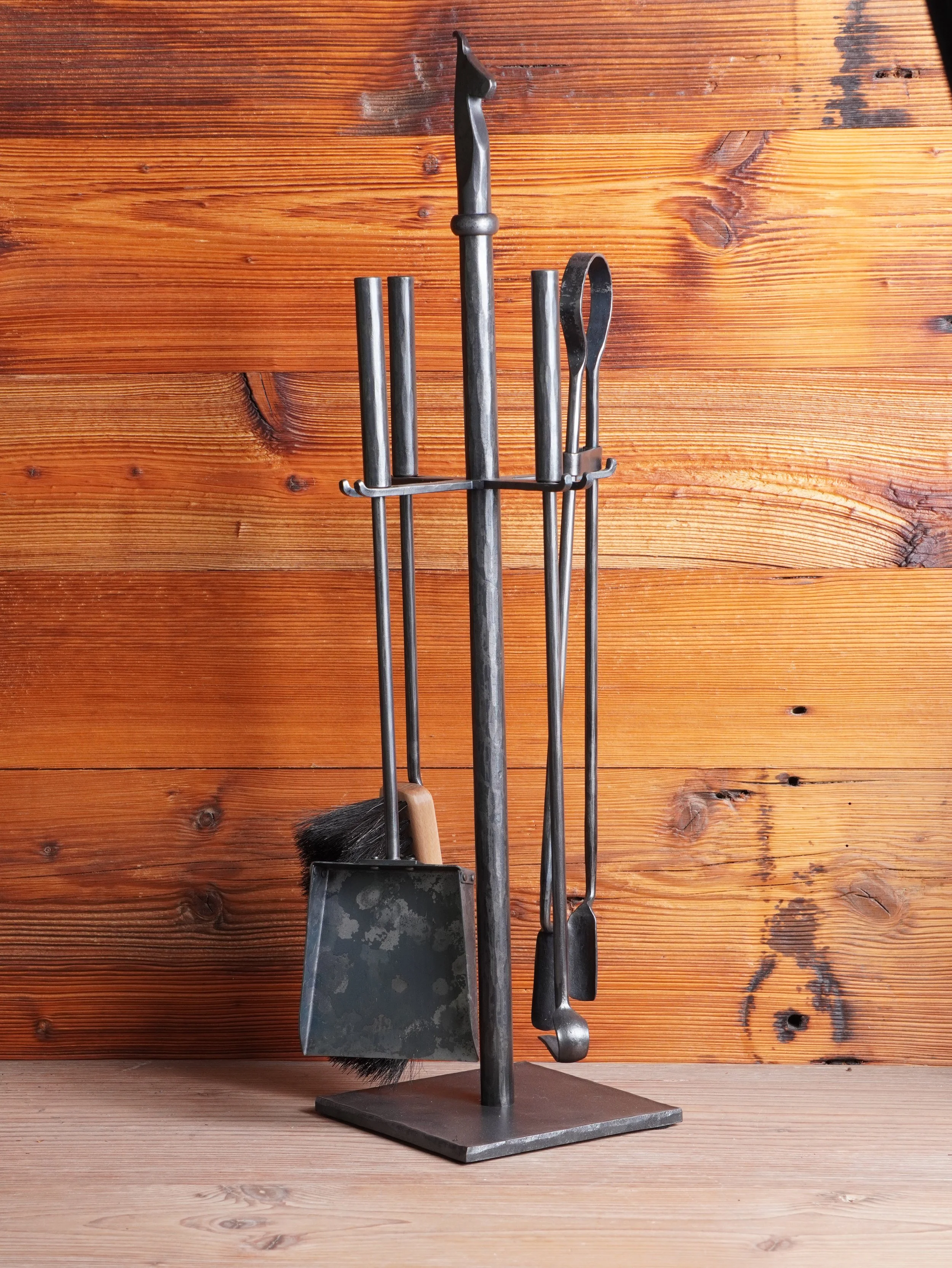 Ständer mit Grillwerkzeugen, einschließlich Bürste, Zange und Schaufel, vor einer Holzwand.