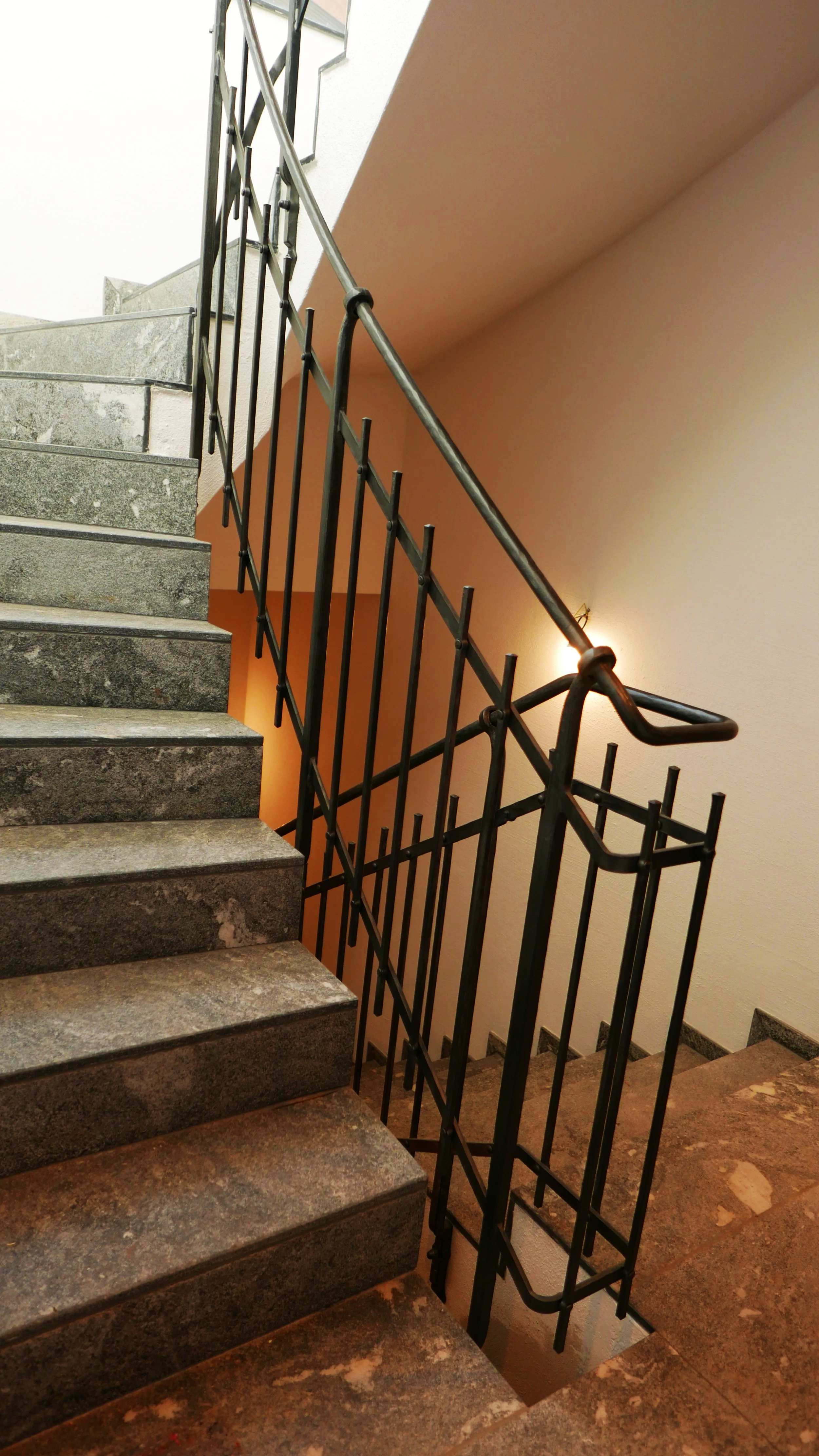 Innentreppenhaus mit grauen Steinstufen, schwarzem Metallgeländer und einer Wand mit warmem Licht.