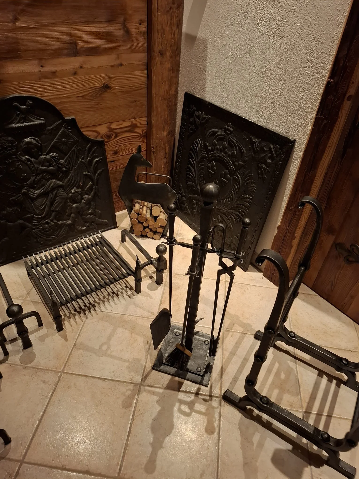 Kaminwerkzeug und Kaminabdeckungen aus Gusseisen, Holzscheite in einem Silobehälter, antike Möbel und Holzverzierungen vor einer Steinwand.
