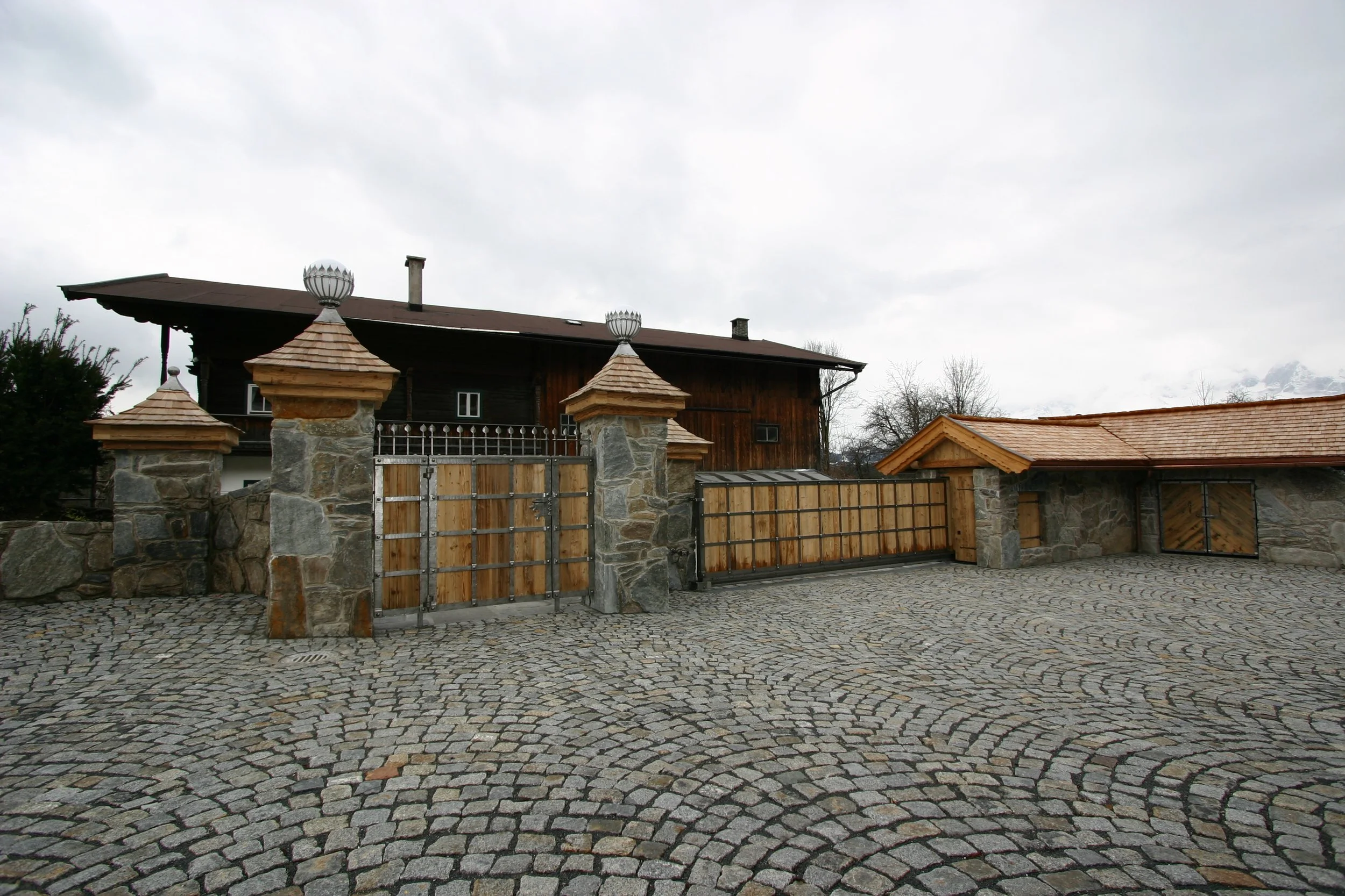 Ein Holztor und ein Zaun vor einem Bauernhaus, umgeben von einem gepflasterten Hof, bei bewölktem Himmel