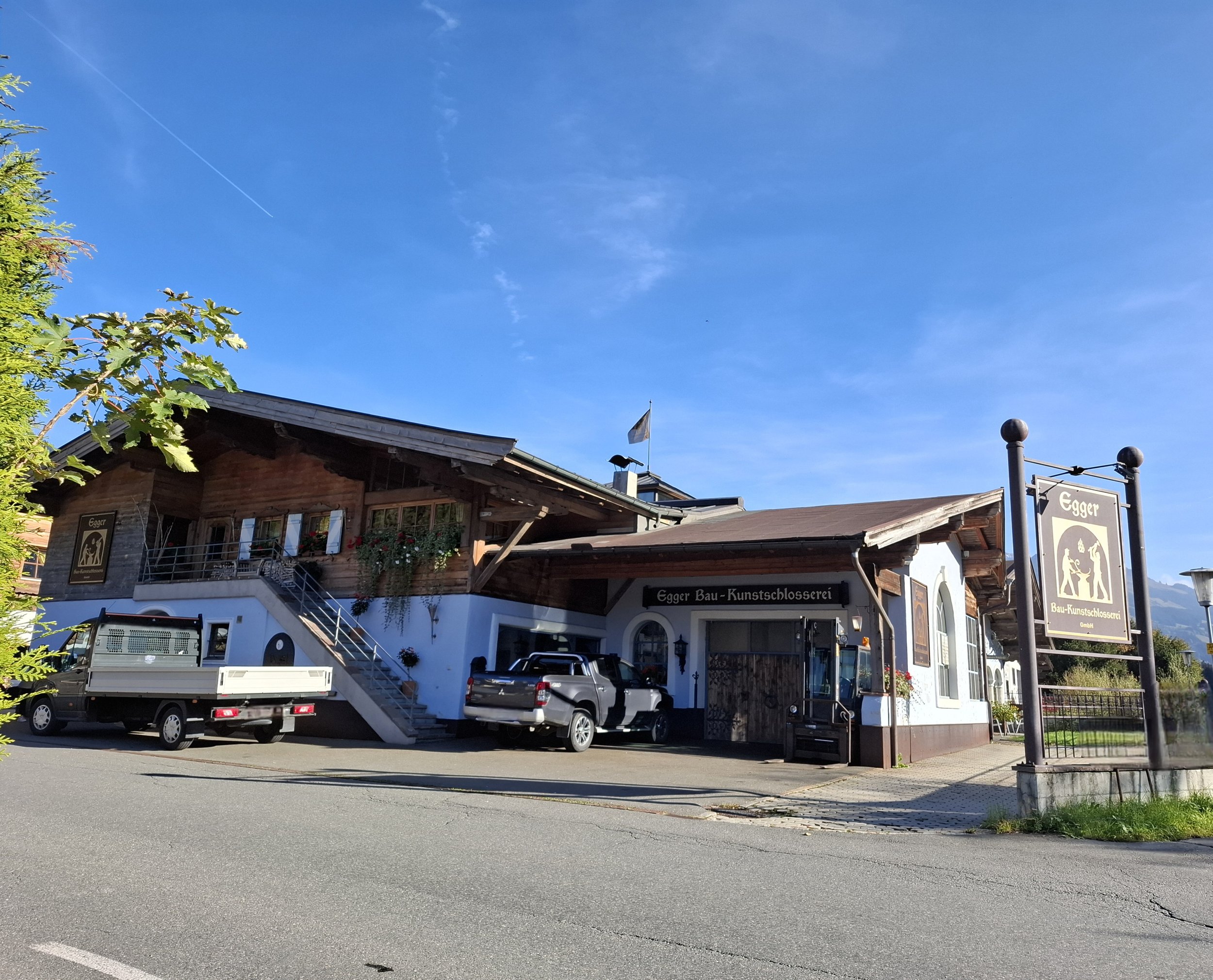 Verwaltung eines Holzhauses mit Anbau, Parkplatz mit Autos, Schilder mit Logo und Name 'Egger Bau-Kunstschlosserei', blauer Himmel, grüner Baum auf der linken Seite.
