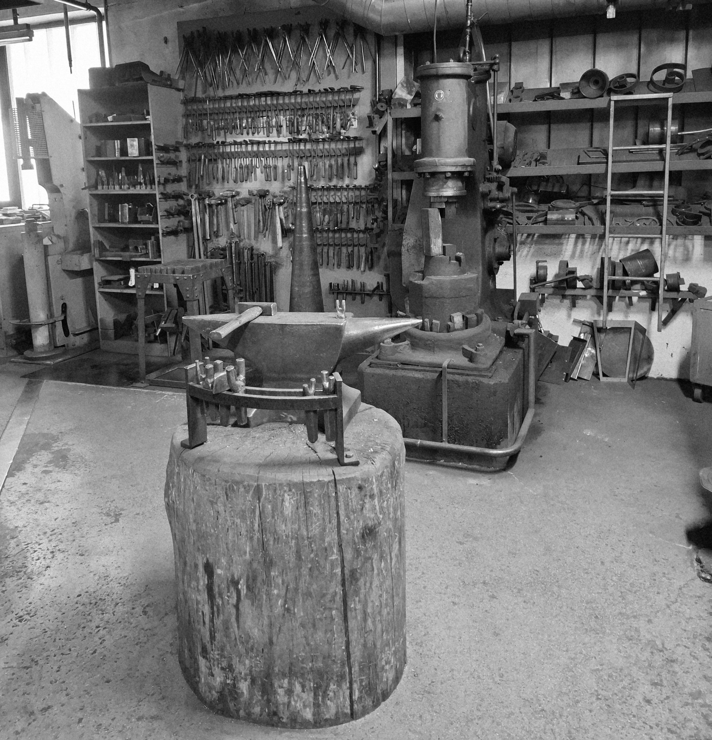 Ein Werkstatt mit verschiedenen Werkzeugen an der Wand und Maschinen im Hintergrund. Im Vordergrund ein Amboss auf einem Baumstamm.