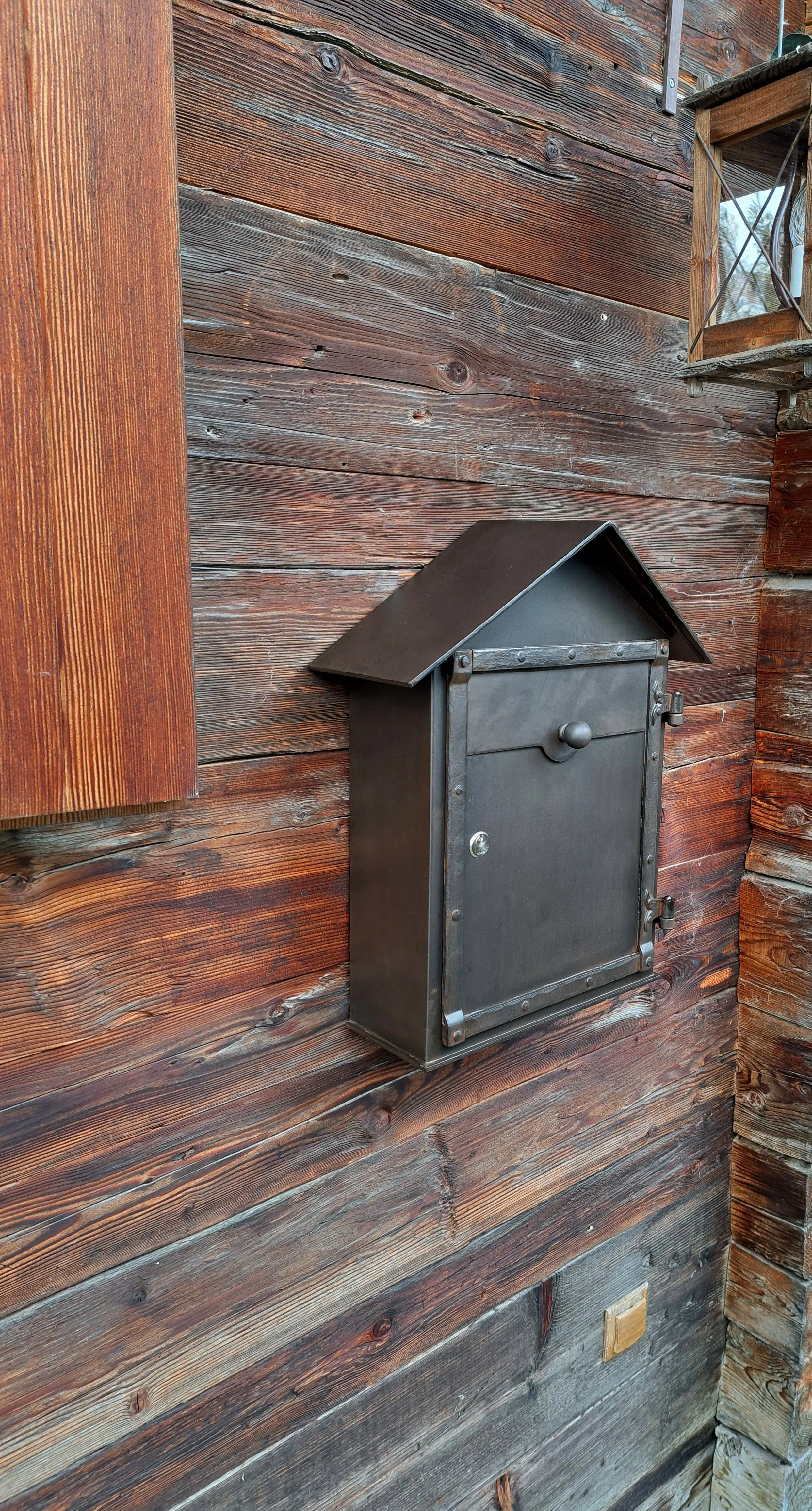 Ein schwarzer Briefkasten an einer Holzwand, der ein Dach hat.