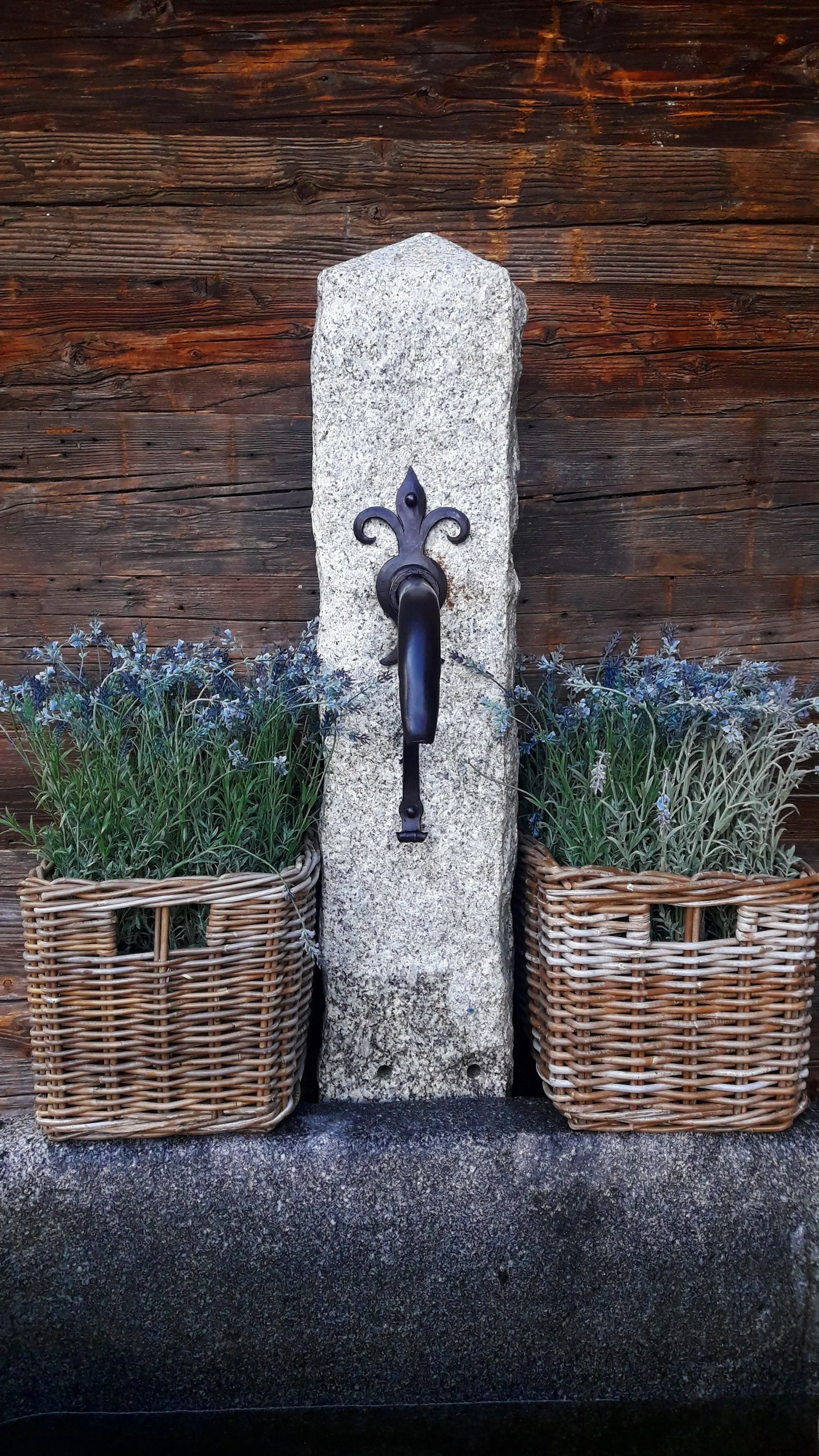 Ein Wasserhahn, der an einem Stein montiert ist, zwischen zwei Korb-Pflanzen mit blauen Blumen, vor einer Holzverkleidung.