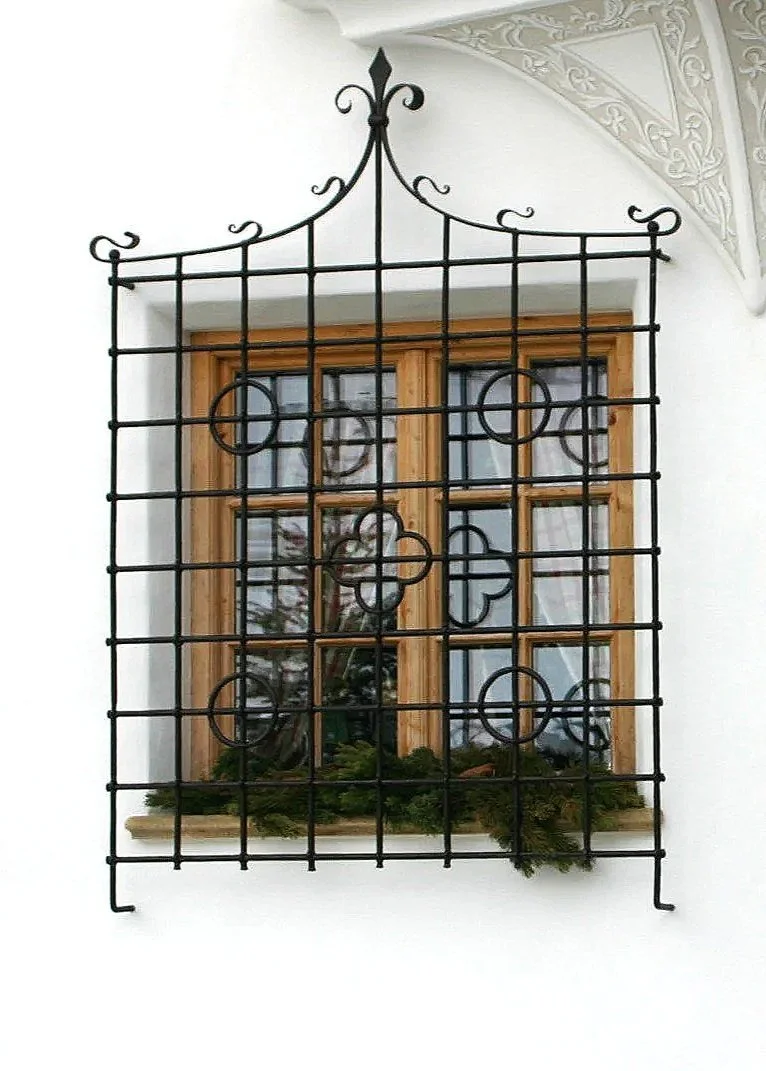Fenster mit Holzrahmen und Gitter vor einer weißen Wand, Dekor in der oberen Ecke, mit Pflanzen auf der Fensterbank.