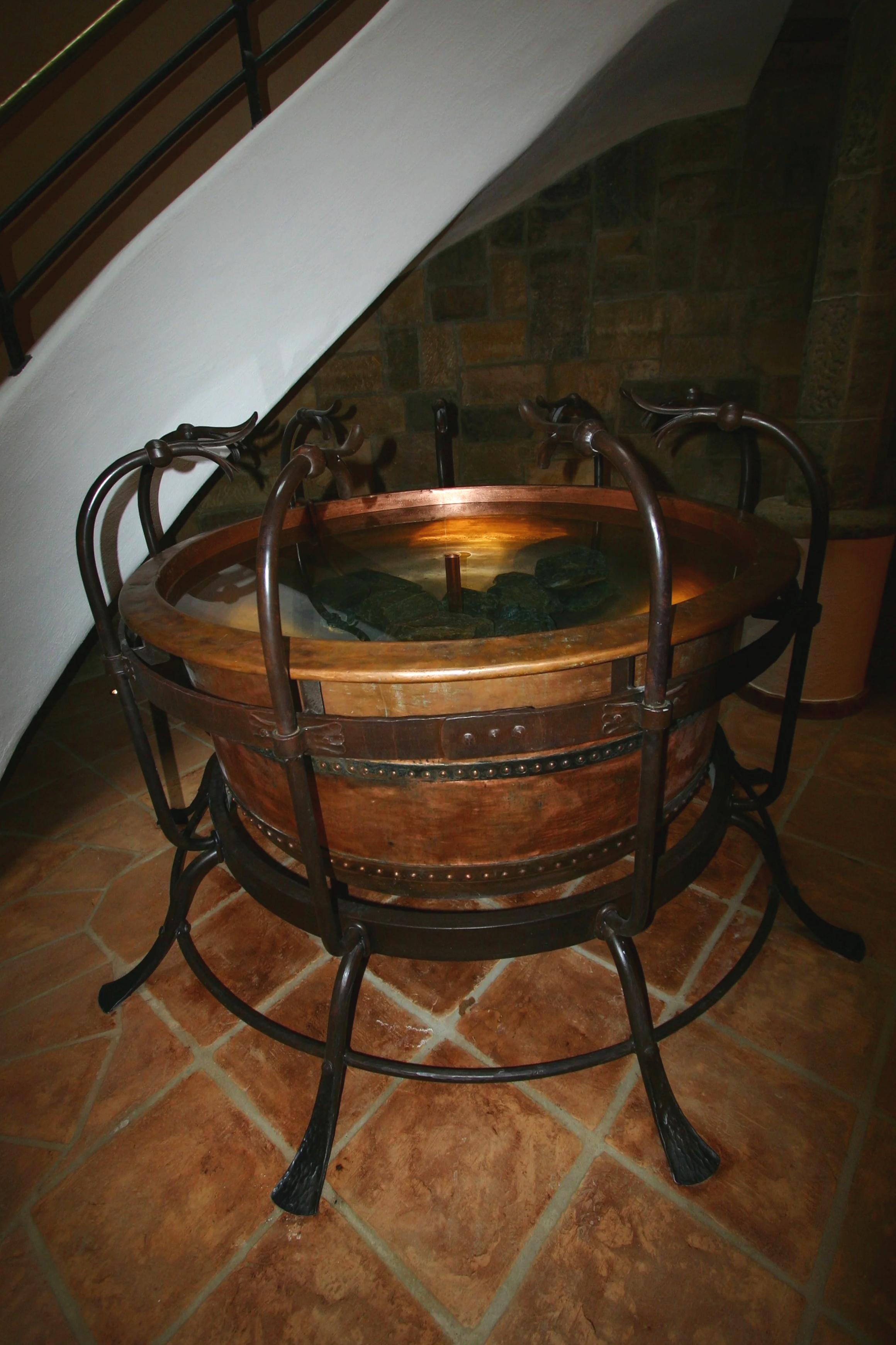 Ein runder, antiker Wassertrog aus Holz mit schwarzen Steinen und Wasser, umgeben von einem schmiedeeisernen Gestell, im Innenbereich auf Terrakottafliesen neben einer weißen Wand und einer Holztreppe.
