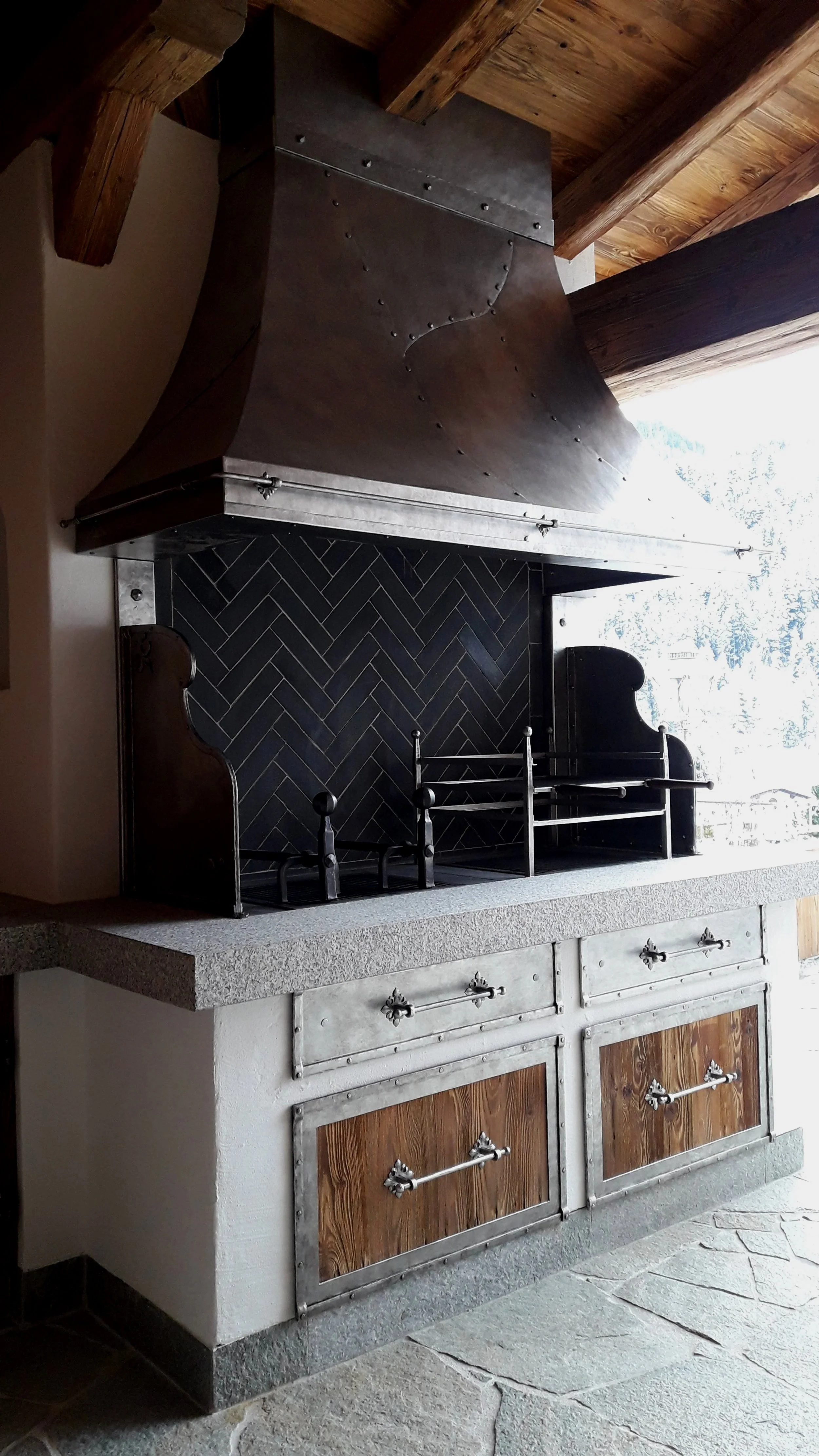 Traditioneller Holzofen mit schwarzer Backstein-Ofenverkleidung und Metallhaube, auf Granitplatte. Holz- und Steinsockel darunter. Blick nach draußen auf verschneite Landschaft.