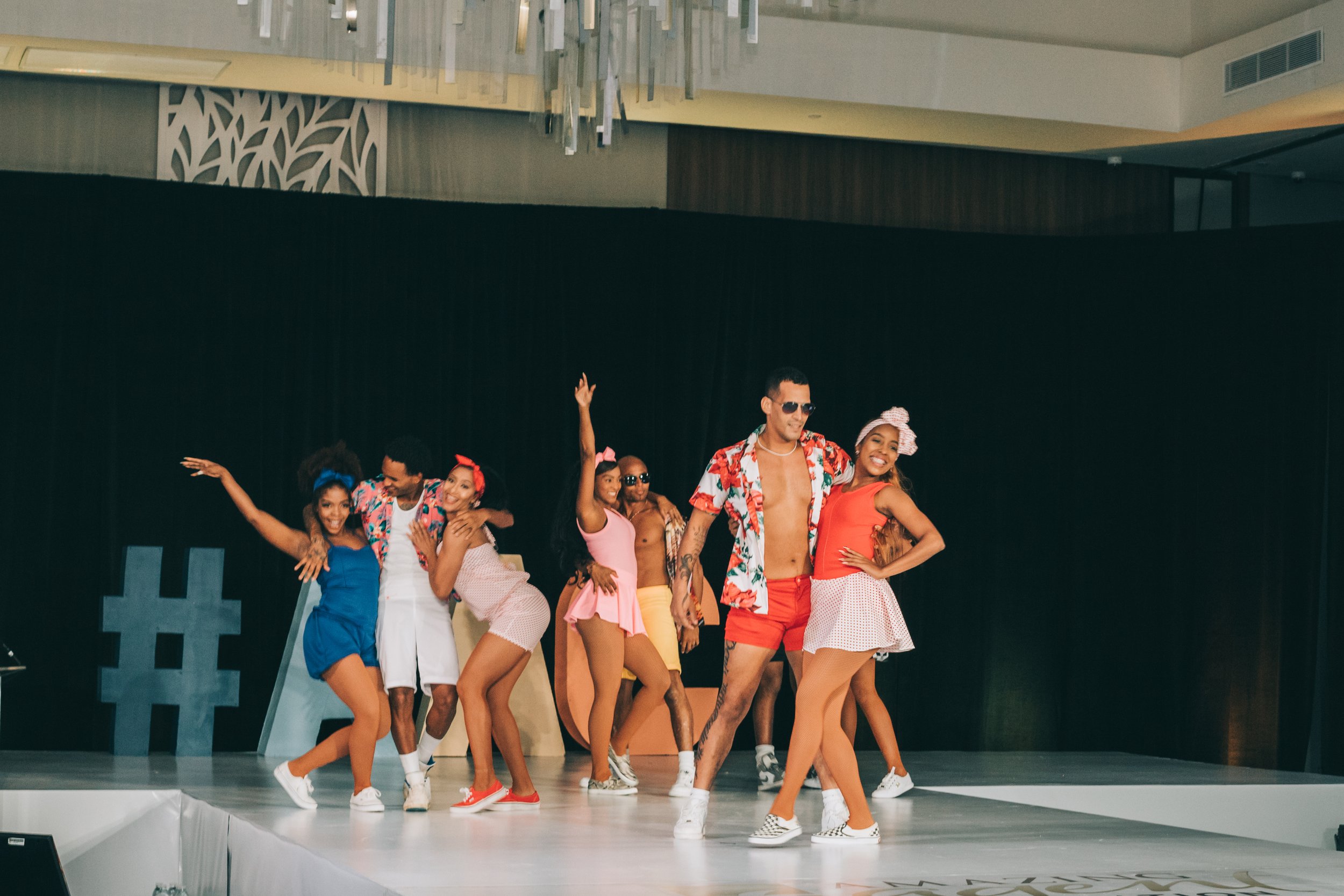 A group of people dressed in colorful, retro-style clothing are dancing on a stage with a black backdrop.
