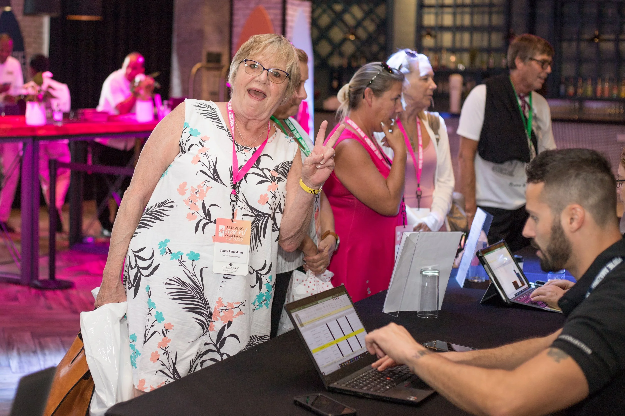 A group of people at a conference or event, with a woman in the foreground making a peace sign and wearing a name tag that says "Sandy Patzkon". Several people are seated at a table with laptops, and the background features a bar area with bottles and glasses.