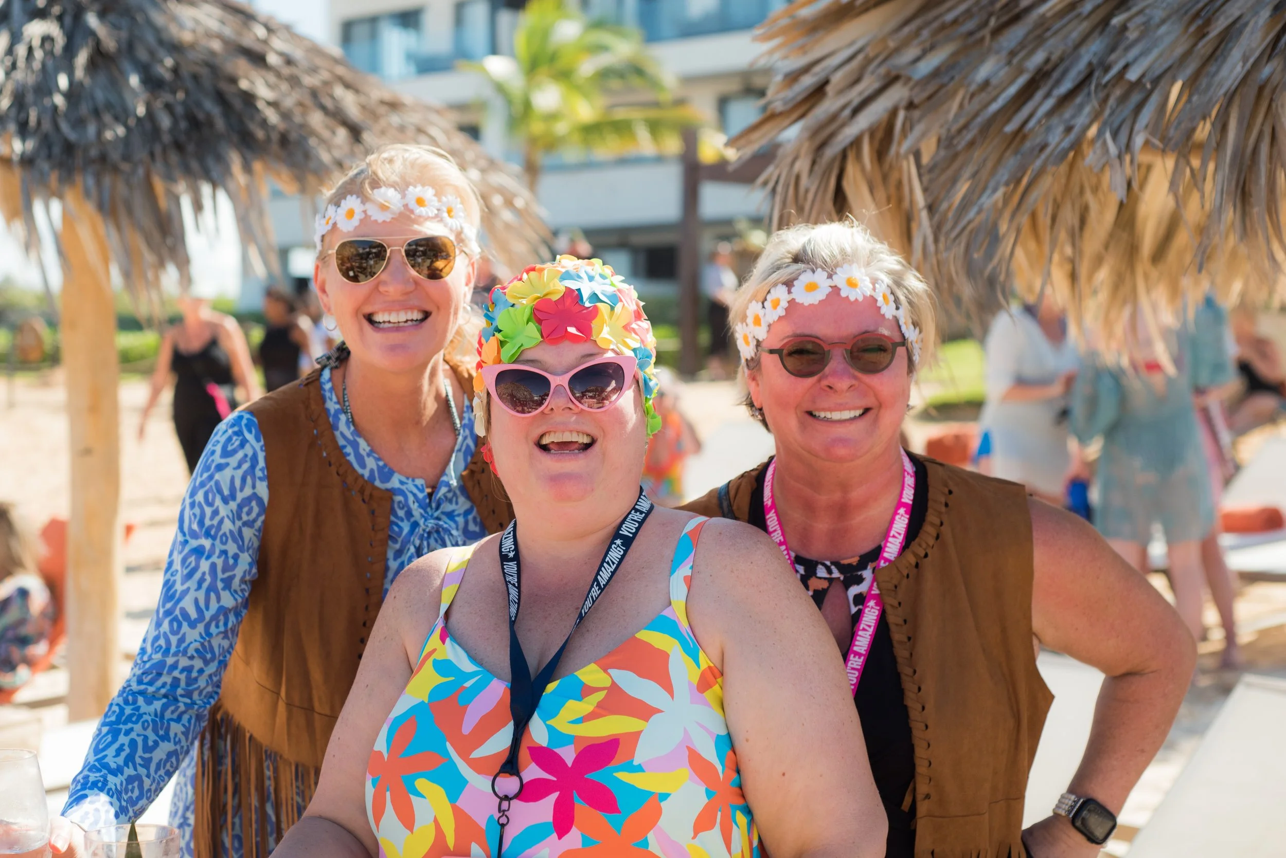 Three women smiling outdoors at a beach or tropical setting, wearing floral headbands and sunglasses, with thatched umbrellas in the background.