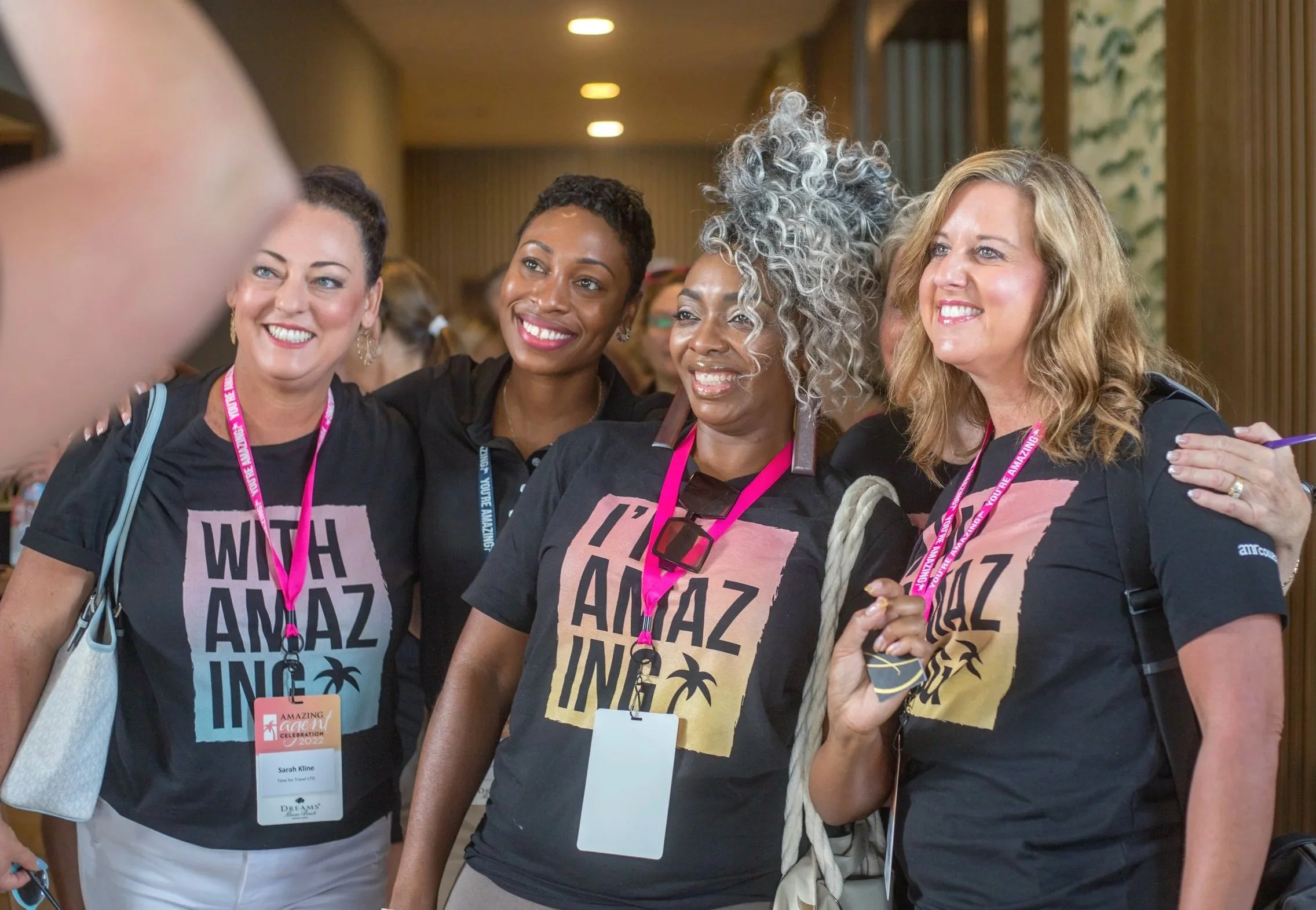 Group of women smiling and taking a selfie at an event, wearing black t-shirts with the words 'WHAT ANAZ IN' and pink lanyards.