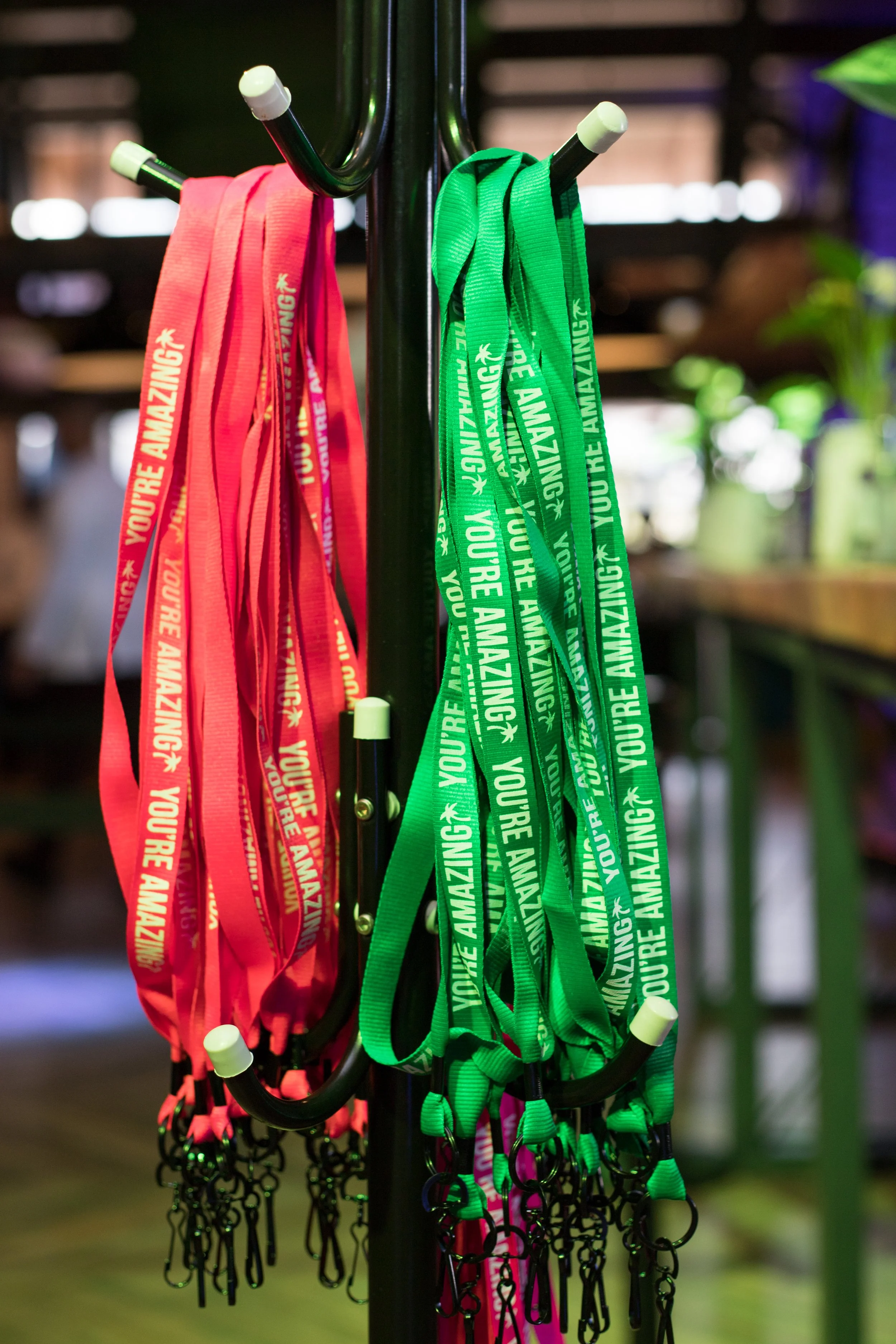 Colorful lanyards with black hooks hanging on a black metal stand, with a blurred indoor background. The lanyards are red, pink, green, and purple, with white text that reads 'You're Amazing' and a small star icon.