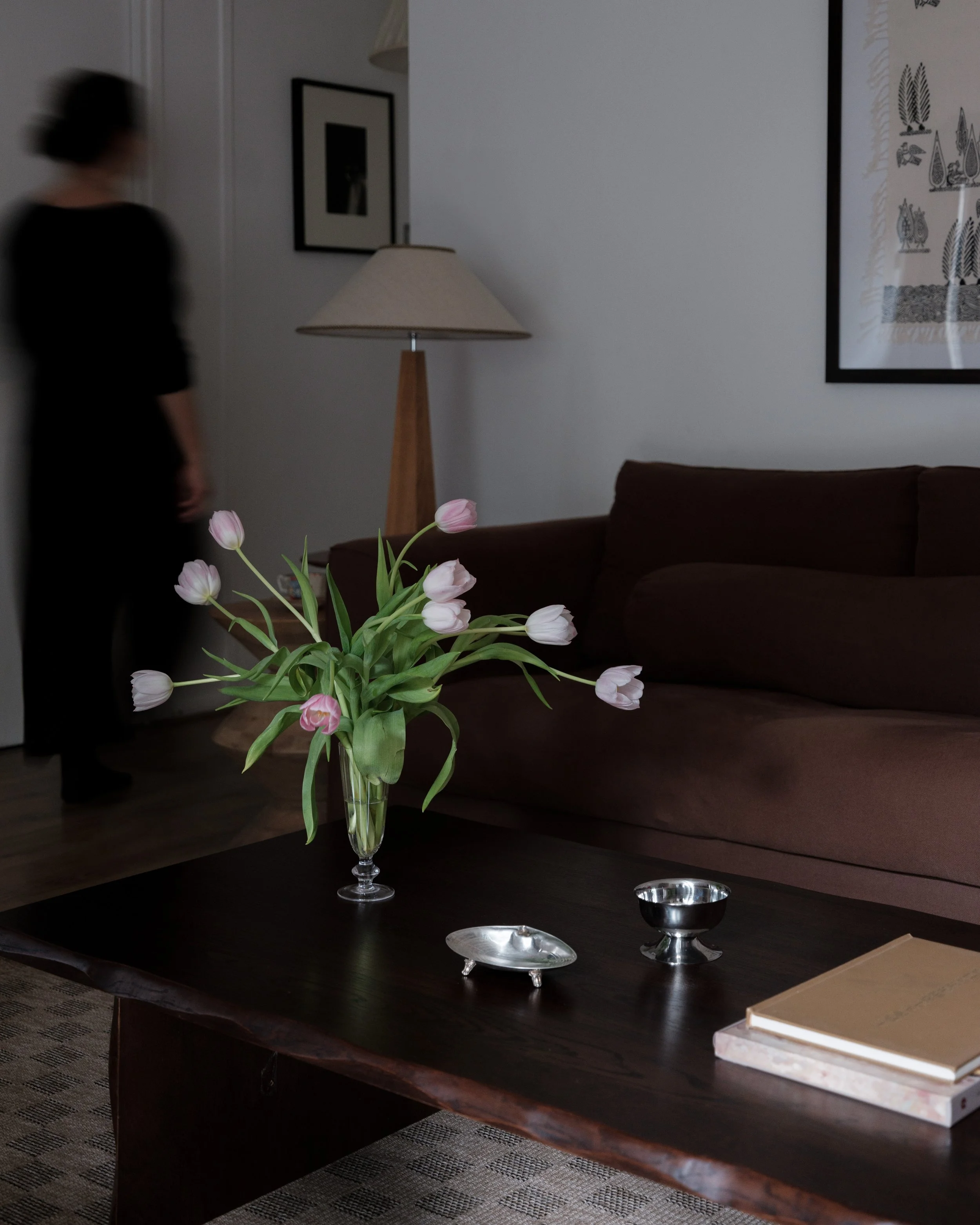 Living room with a dark brown couch, a wooden coffee table with a glass vase of pink and white tulips, a small silver bowl, a book, a floor lamp, and framed artwork on the wall. A person is blurred in the background.