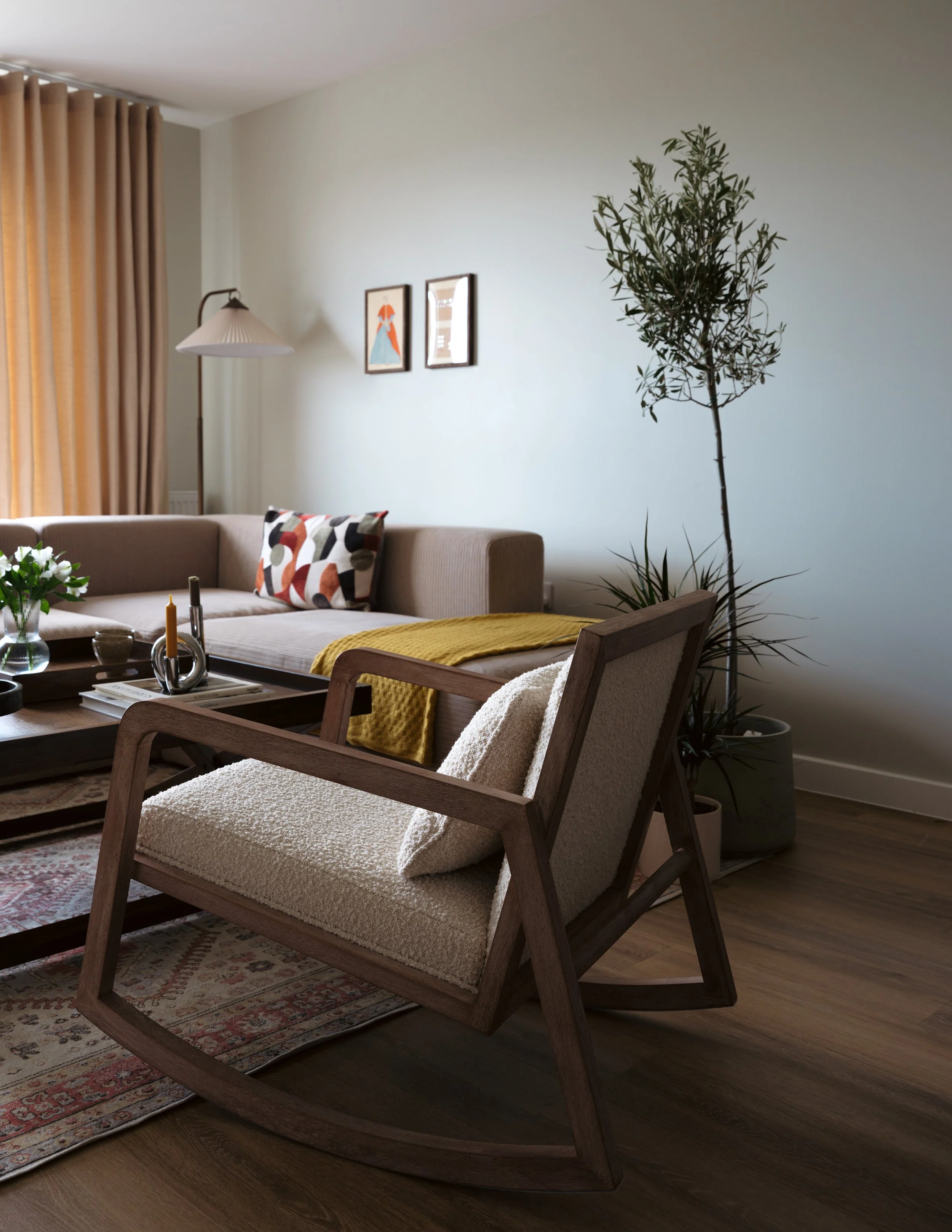 Living room with beige sofa, wooden chair with cream cushion, large potted plant, artwork on wall, and window with beige curtains.