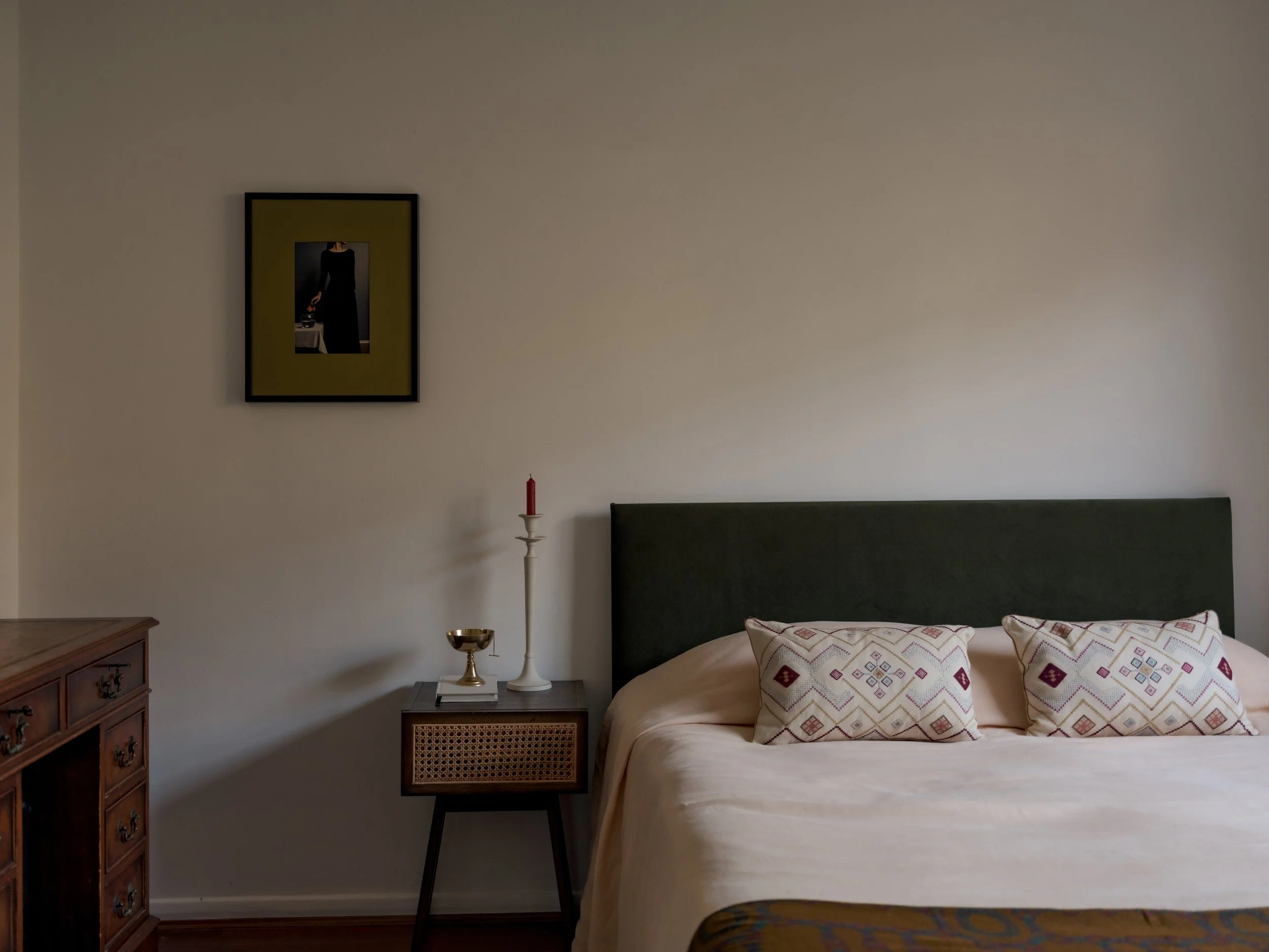 A bedroom with a white wall, a bed with a green headboard and patterned pillows, a wooden nightstand with a candle, a silver bowl, and a book, and a framed photograph of a woman in a black dress hanging above.