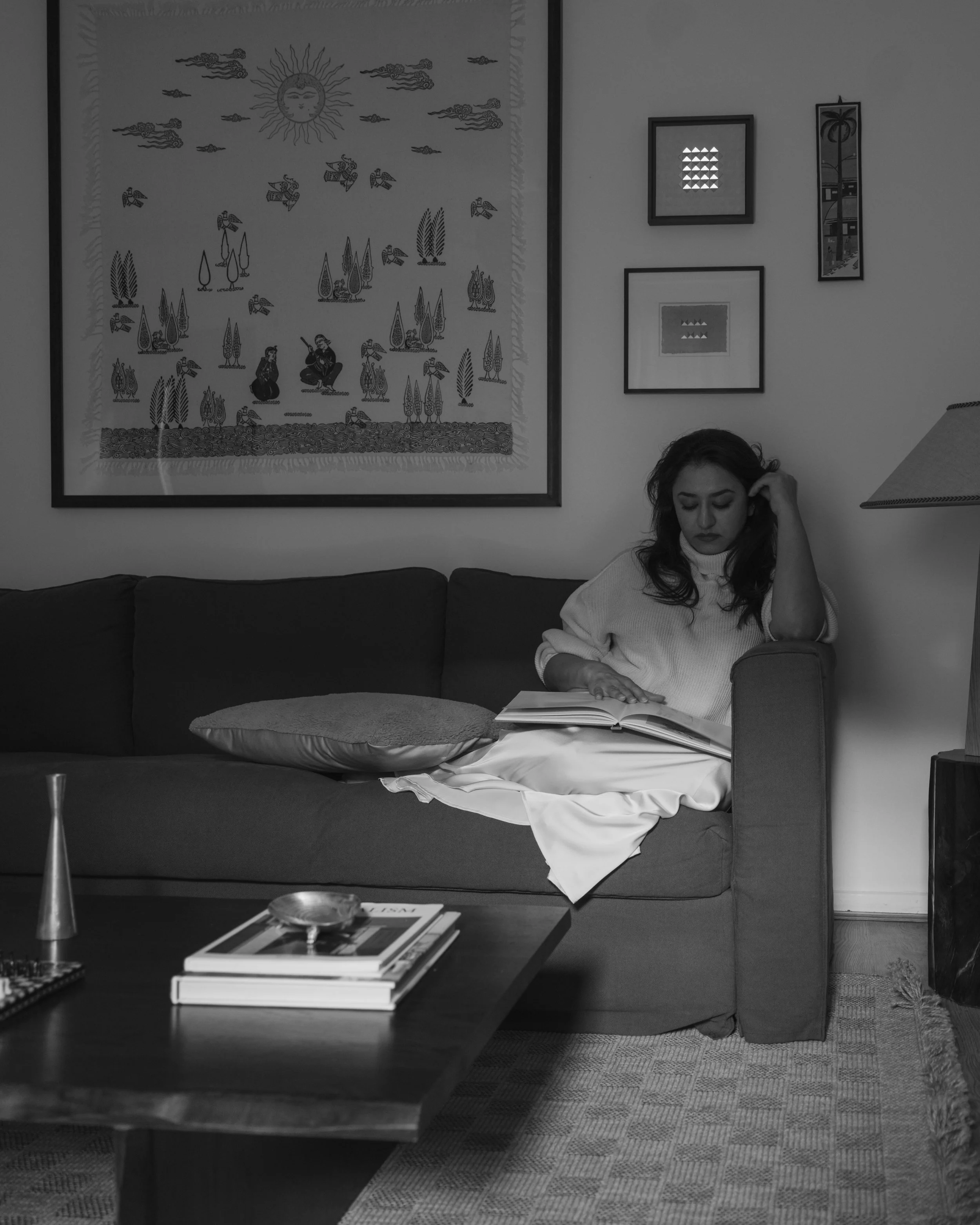 A woman sitting on a sofa reading a book in a room with framed artwork on the wall, a table with books and a decorative item in the foreground, and a lamp beside her.