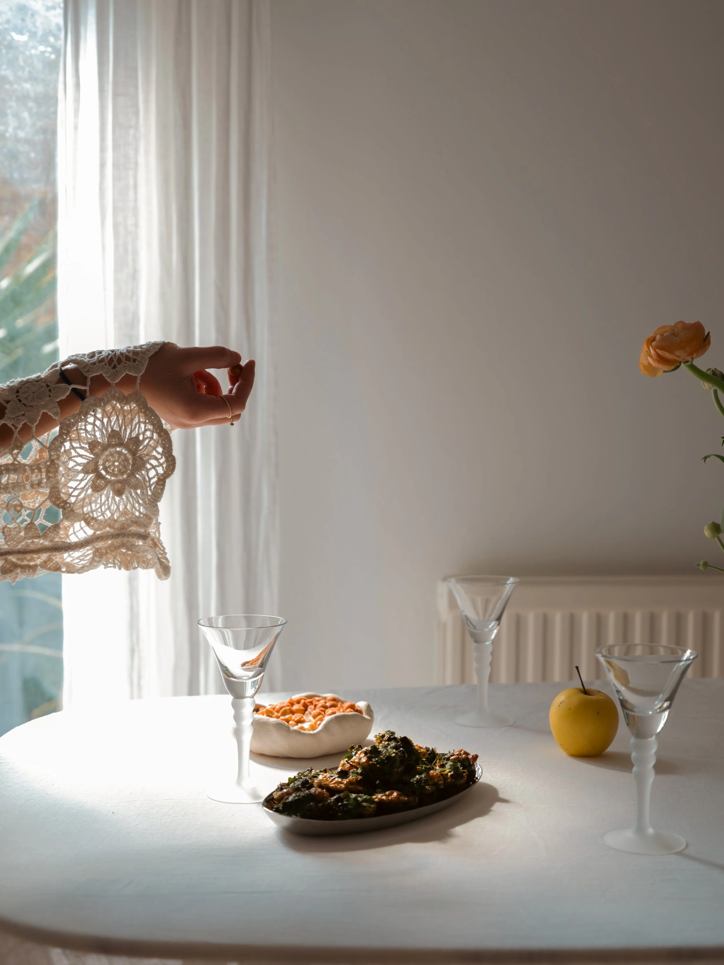 A table set with three empty glasses, a yellow apple, and two dishes of food, with a person's hand reaching out near the left side, wearing a lace sleeve. The background has a window with white curtains and some greenery outside.