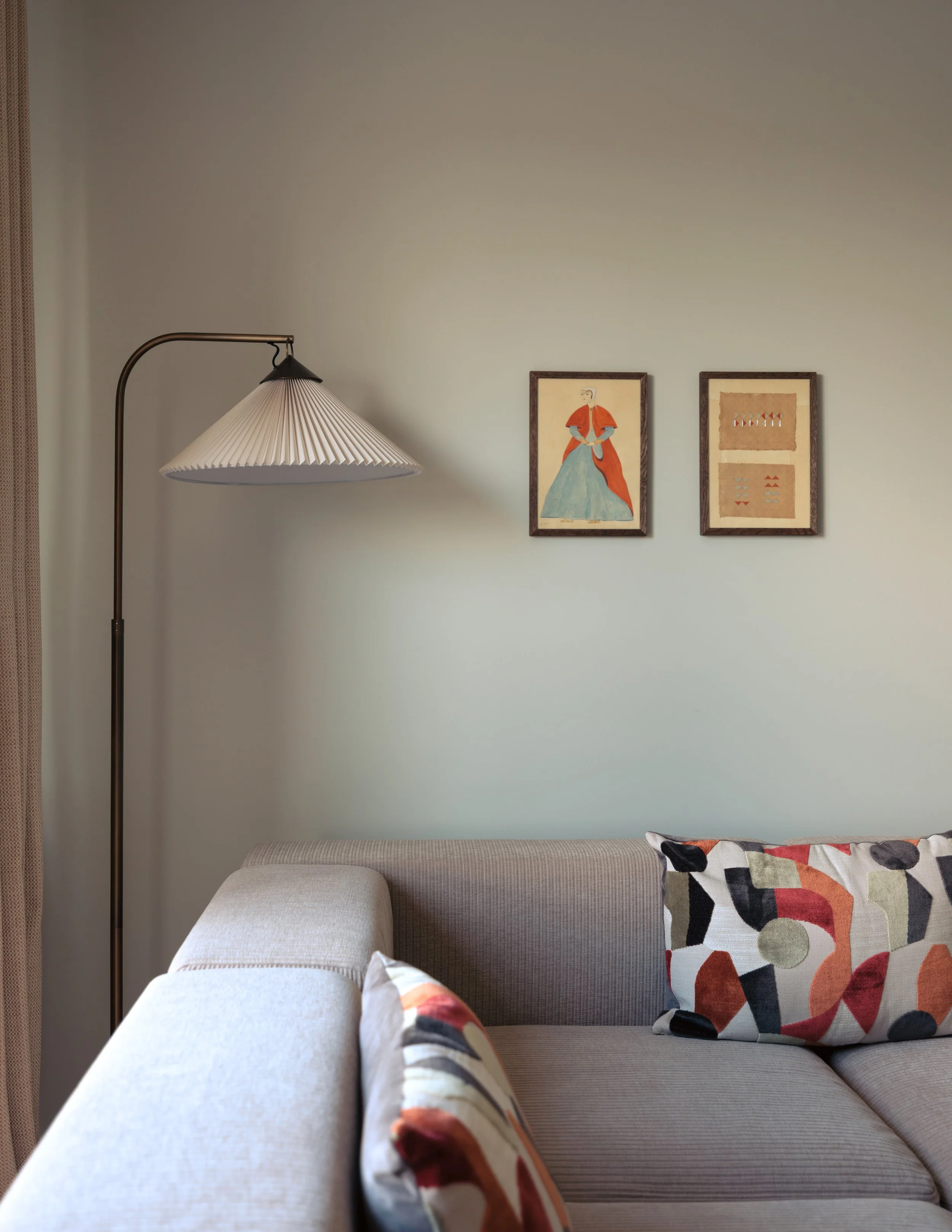 Living room with beige sectional sofa, two decorative pillows with abstract patterns, a standing floor lamp with a pleated shade, and two framed art pieces on a light-colored wall.