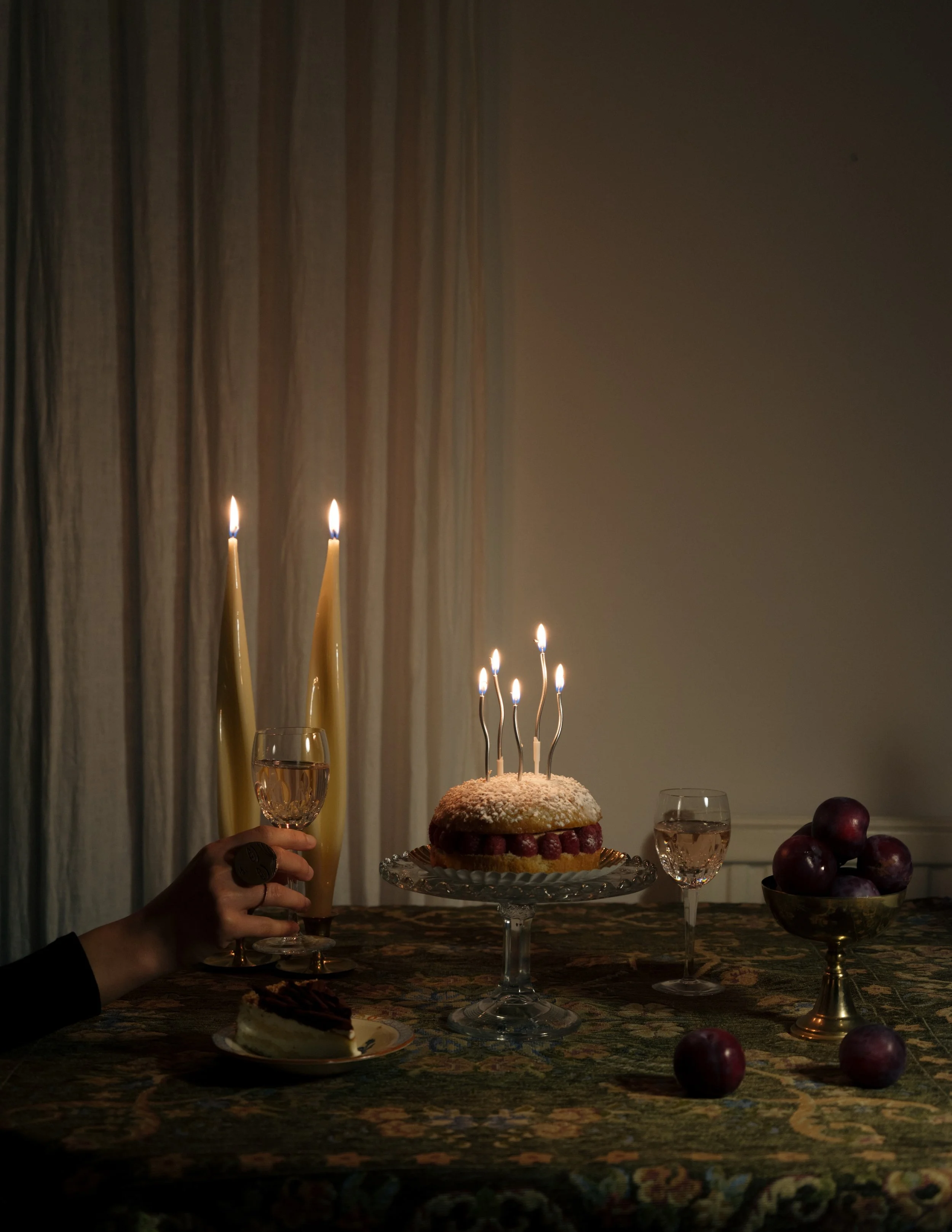 A birthday table setting with a cake with lit candles, two glasses of water, a slice of cake on a plate, a bowl of red apples, and three tall yellow candles in the background.