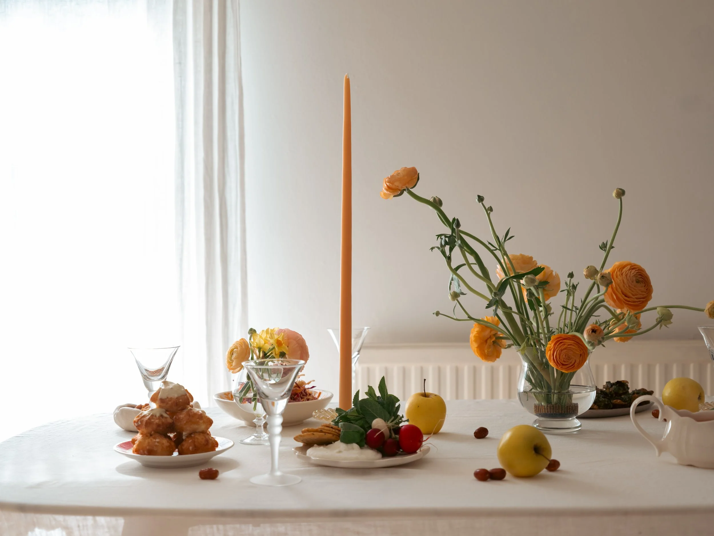 Table set with flowers, apples, a lit candle, and food, with a curtain and window in the background.
