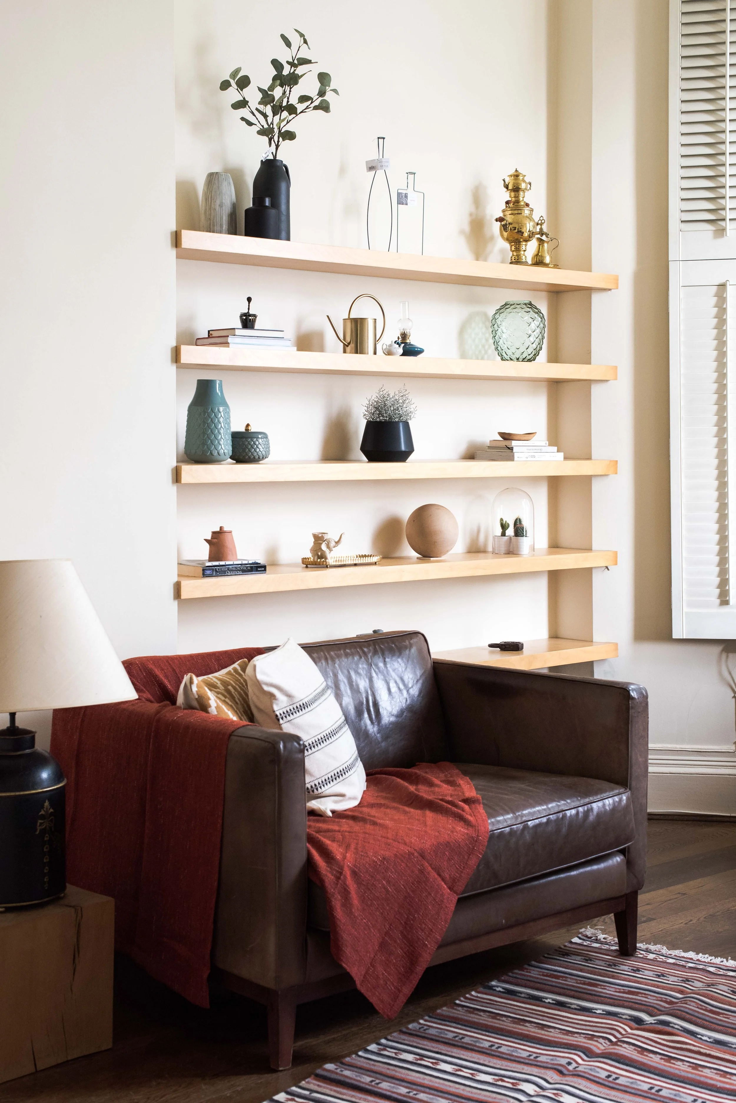 Living room with a brown leather sofa with a red blanket and white and striped pillows. Left of the sofa is a side table with a lamp and a black decorative jar. Behind the sofa, there are four wooden shelves mounted on the wall, displaying various va