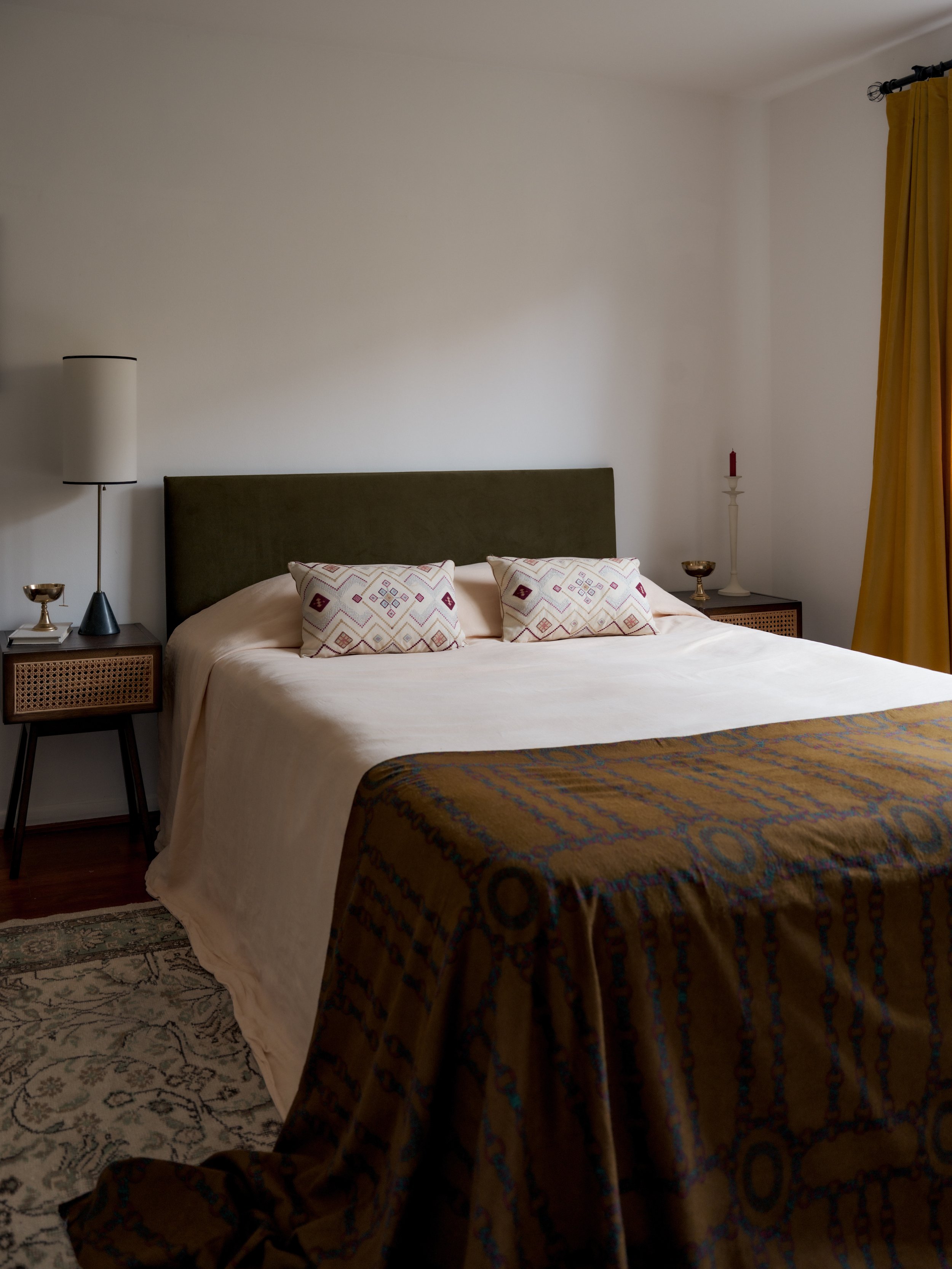 A neatly made bed with a green headboard, decorative pillows, a cream-colored bedspread, and a patterned bedskirt in a bedroom with side tables, lamps, and curtains.