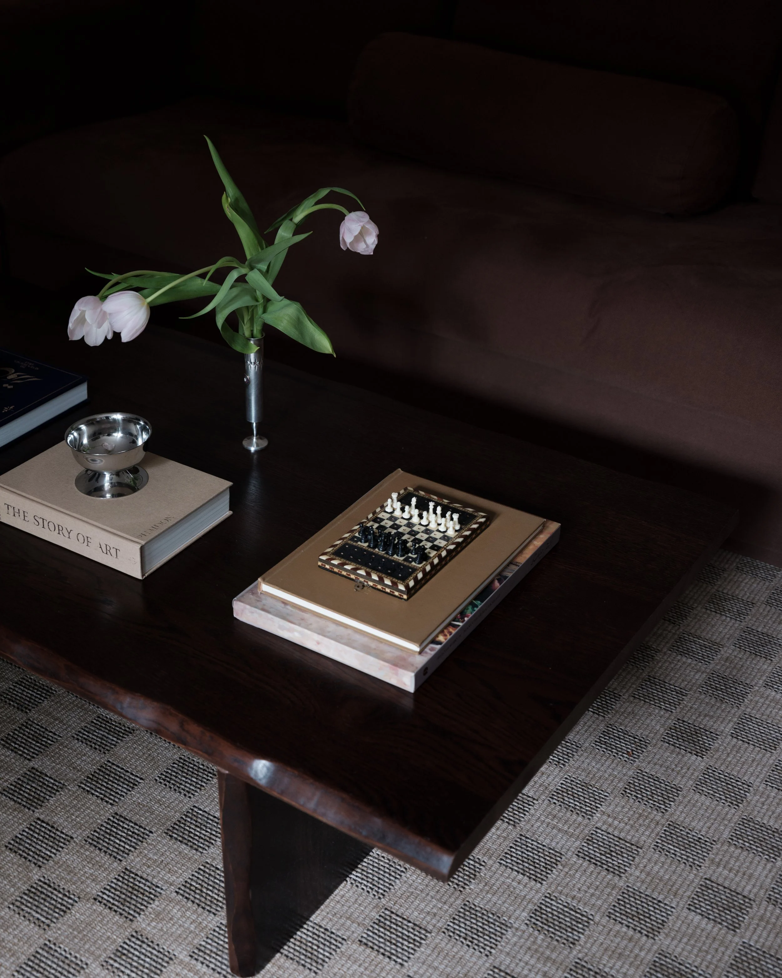 Dark wooden coffee table with a bouquet of white tulips in a glass vase, a silver bowl on top of a book titled 'The Story of Art,' and a chess set on a large beige book, with a brown sofa and patterned rug in the background.
