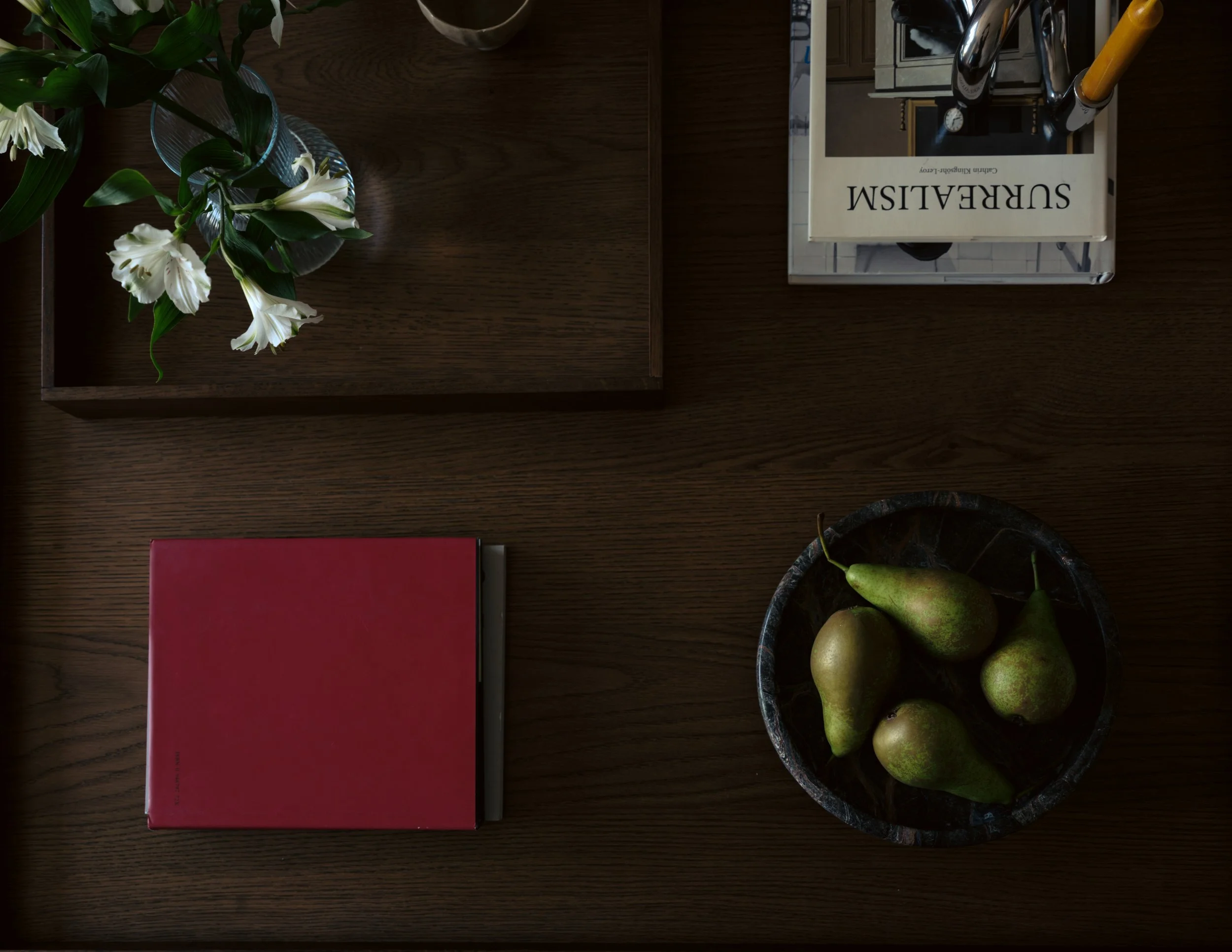 A dark wood table with a flower arrangement of white flowers with green leaves in a clear glass vase, a closed red book, a bowl of green pears, and a box of hairbrushes labeled 'SUREALISM' on top.