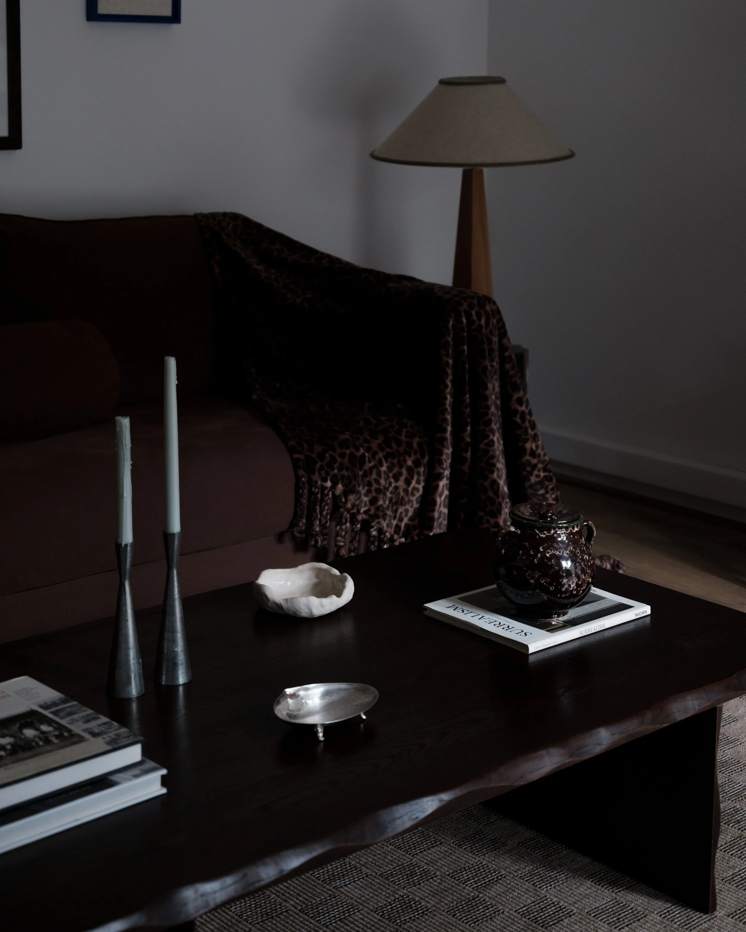 Living room with a dark wooden coffee table, a brown sofa with leopard print throw, a beige floor lamp, and decorative objects including two tall candles, a white bowl, a black vase, and a stack of books.