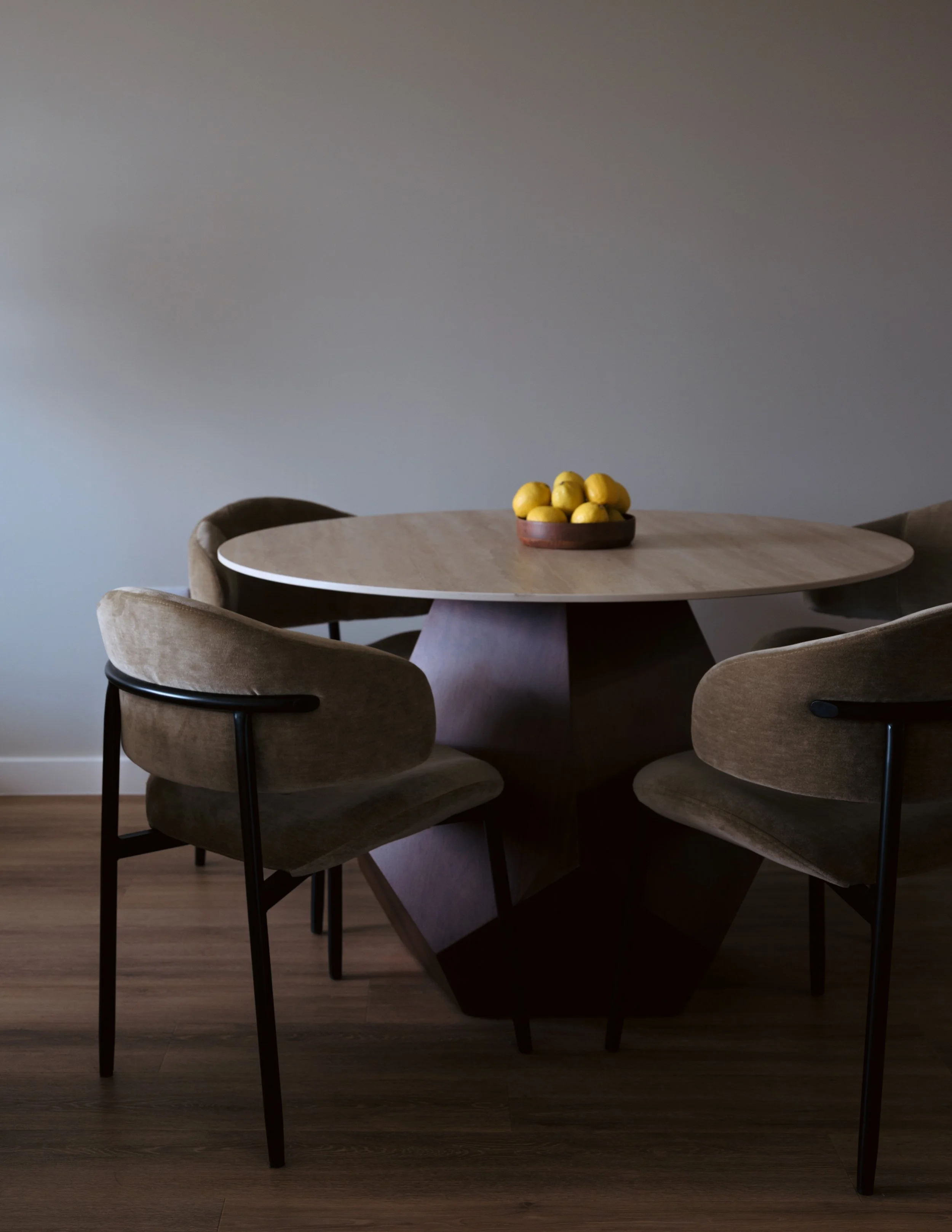 A round wooden dining table with a dark geometric base, four upholstered chairs with black armrests, and a small wooden bowl of yellow lemons on top, set against a plain gray wall.