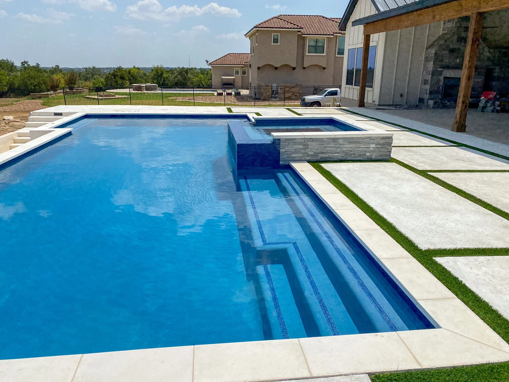 Newly constructed backyard pool with a built-in hot tub, stone accents, concrete paving, and a view of neighboring houses, under a partly cloudy sky.