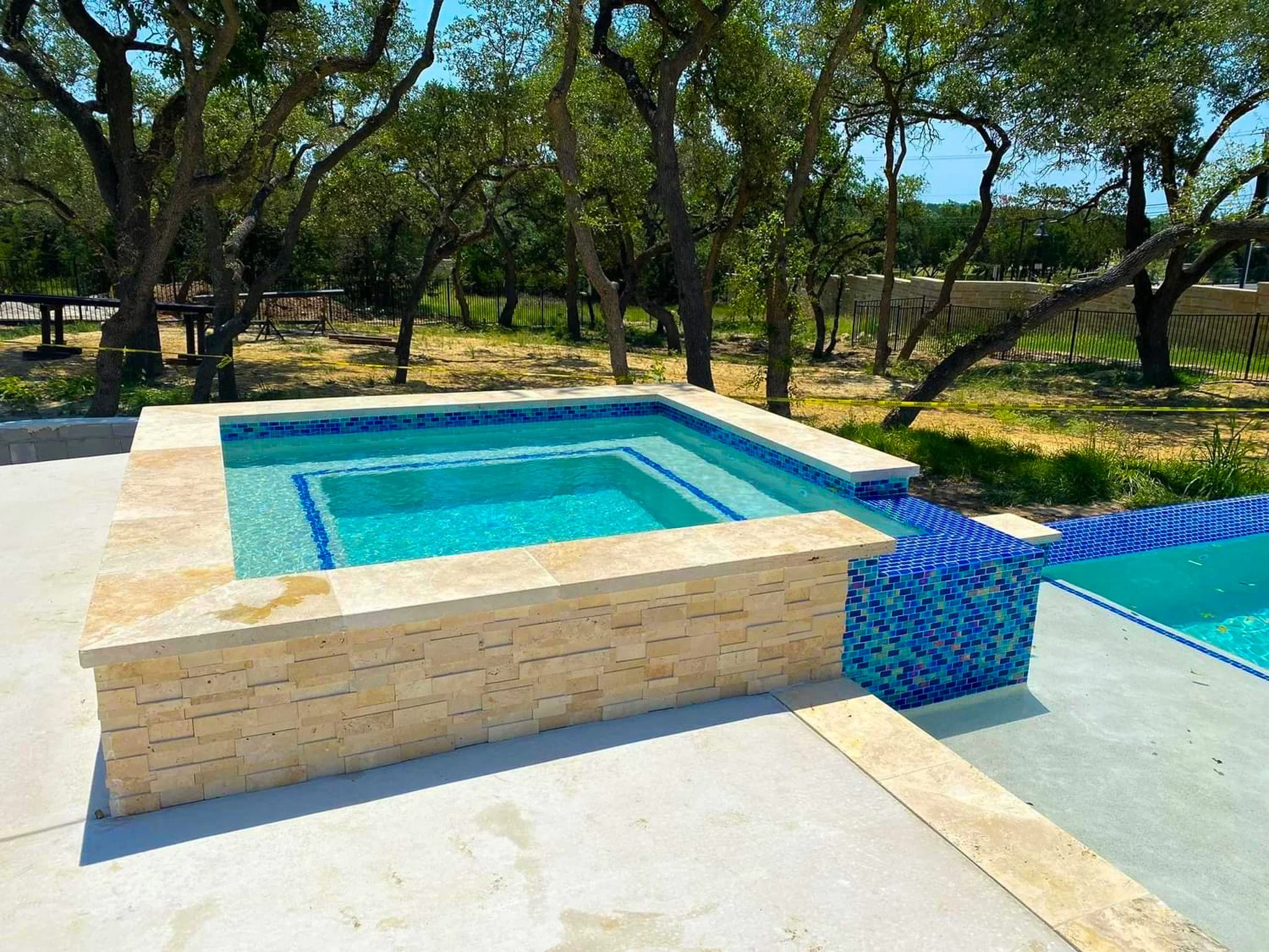 A small rectangular hot tub with a blue tiled water feature on the side, next to a larger pool, in an outdoor area with trees and fencing in the background.