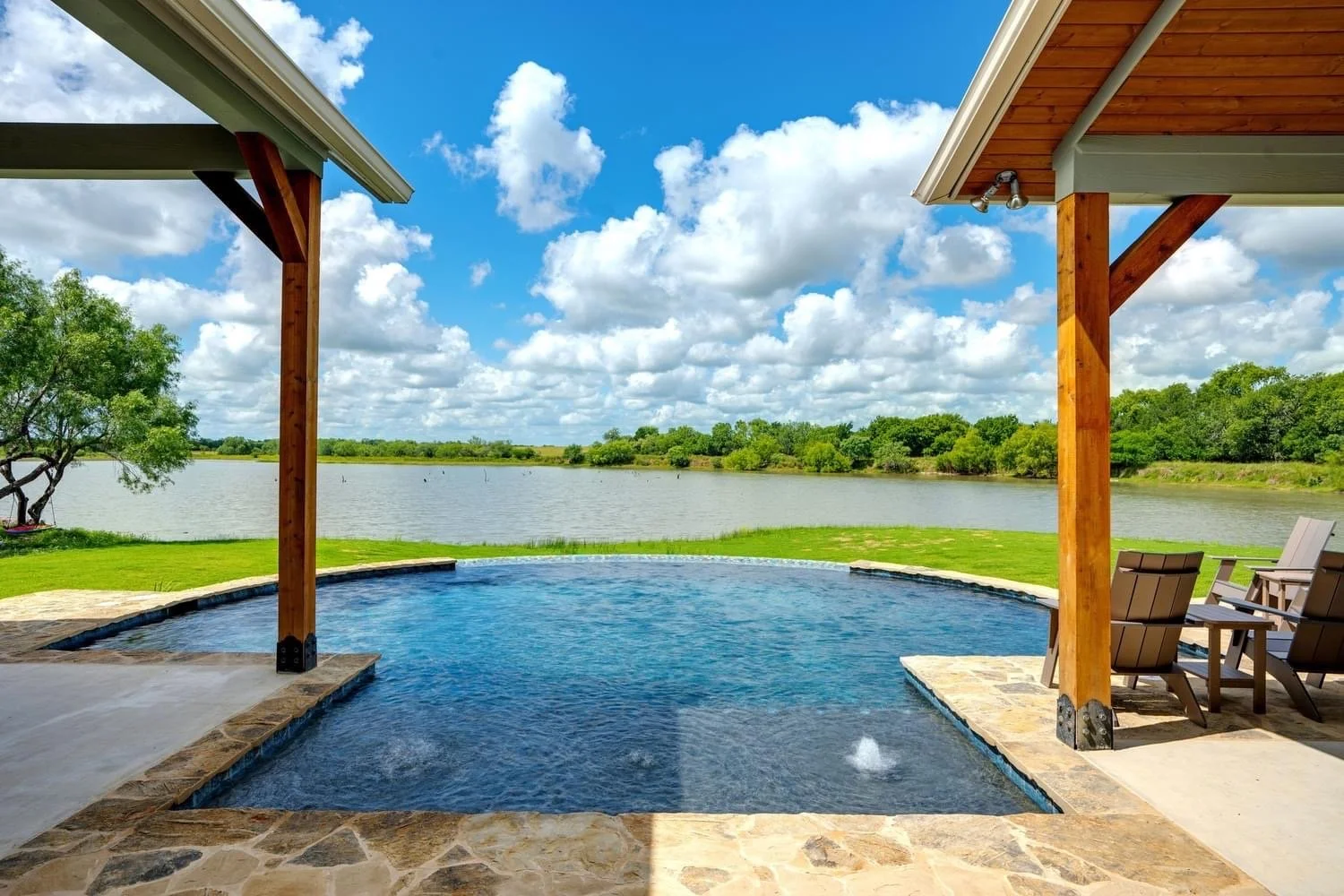 View of a backyard patio with an in-ground pool, overlooking a lake with trees and a blue sky with clouds.