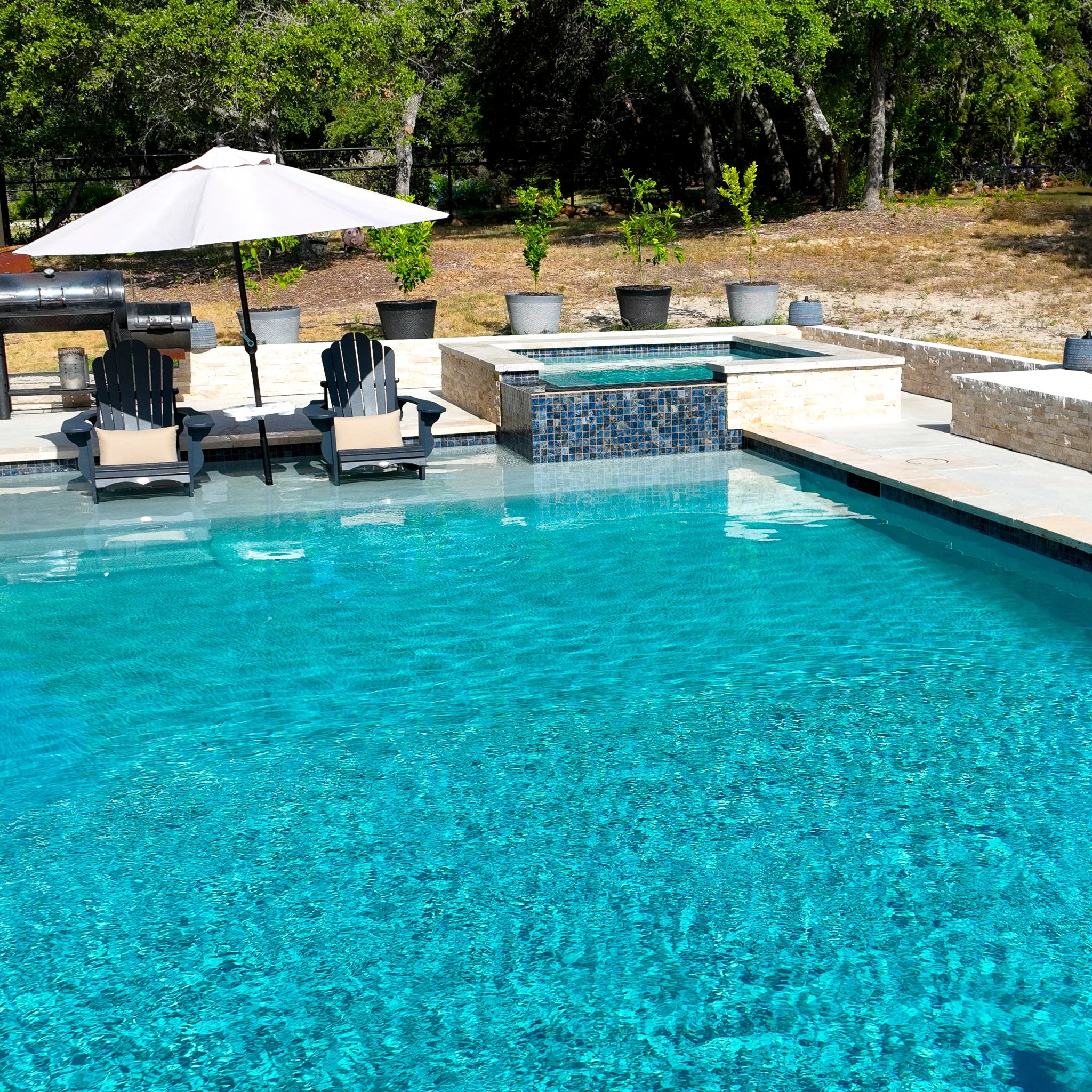A swimming pool with clear blue water, surrounded by a patio with lounge chairs, an umbrella, and potted plants in the background. There are trees behind the patio.