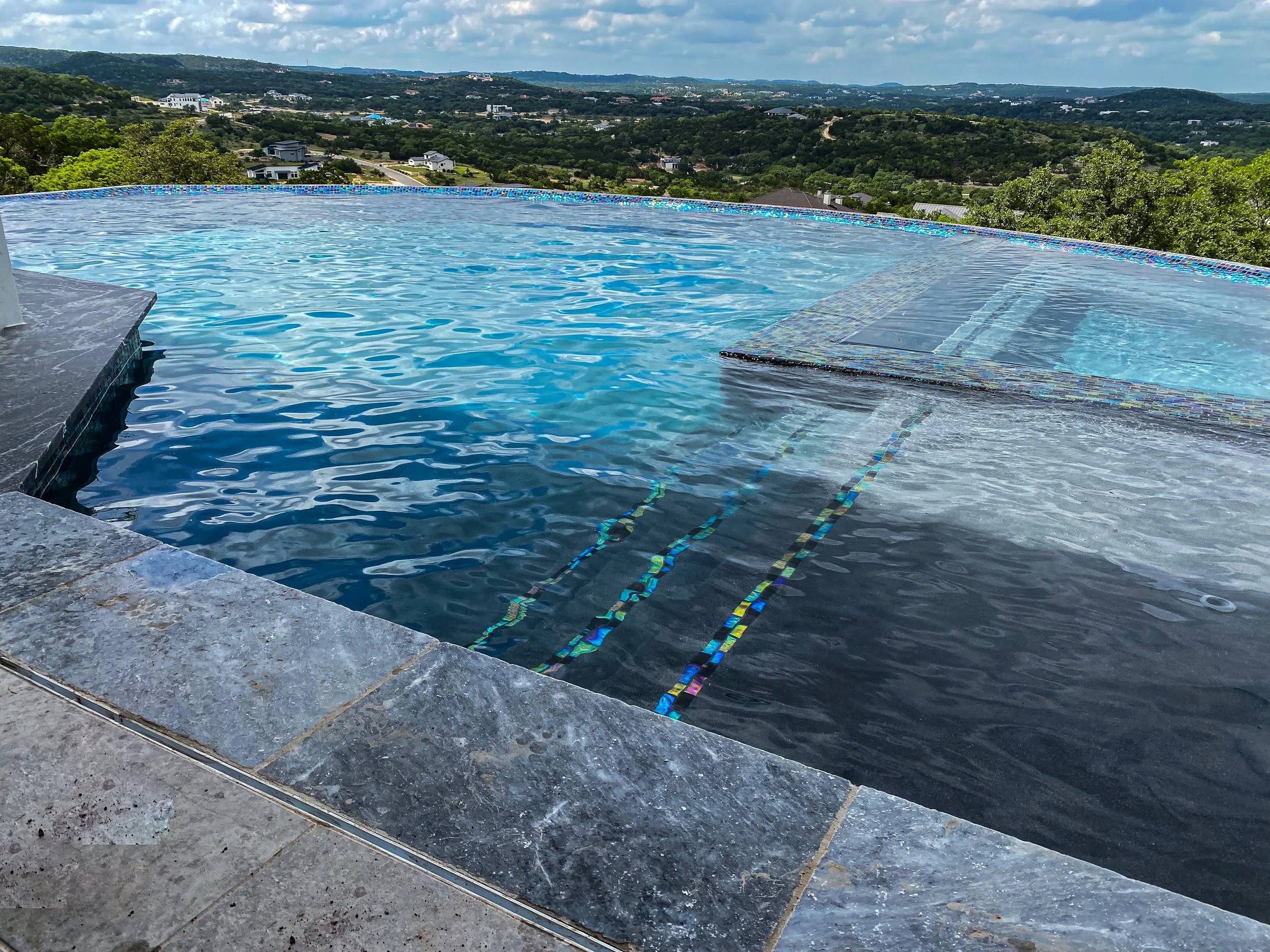 Infinity pool with clear water, tile edge, and a scenic view of green hills and distant houses under a partly cloudy sky.