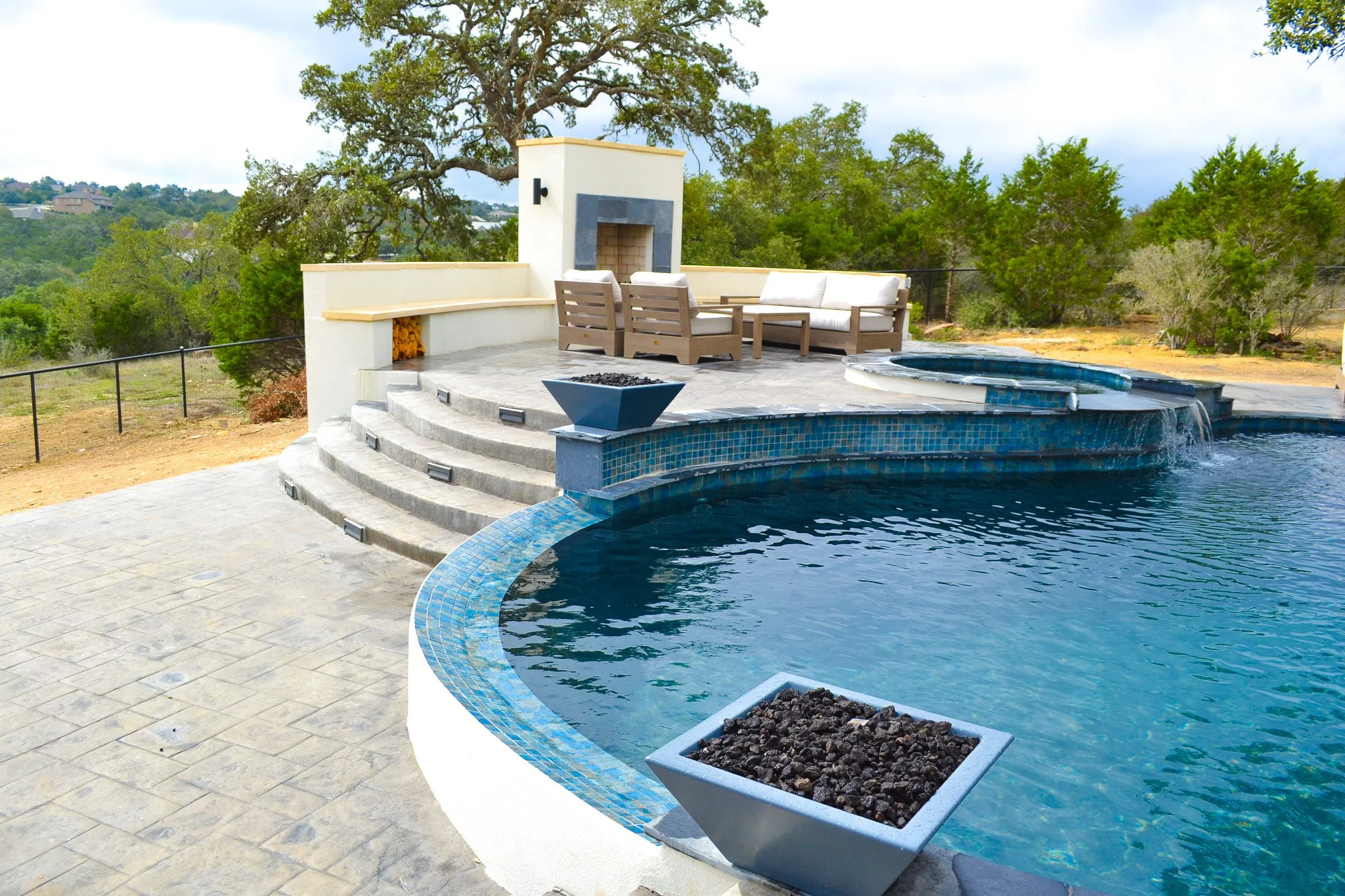 Luxury backyard with a swimming pool, water feature, outdoor seating area, pent fireplace, and lush green trees in the background.