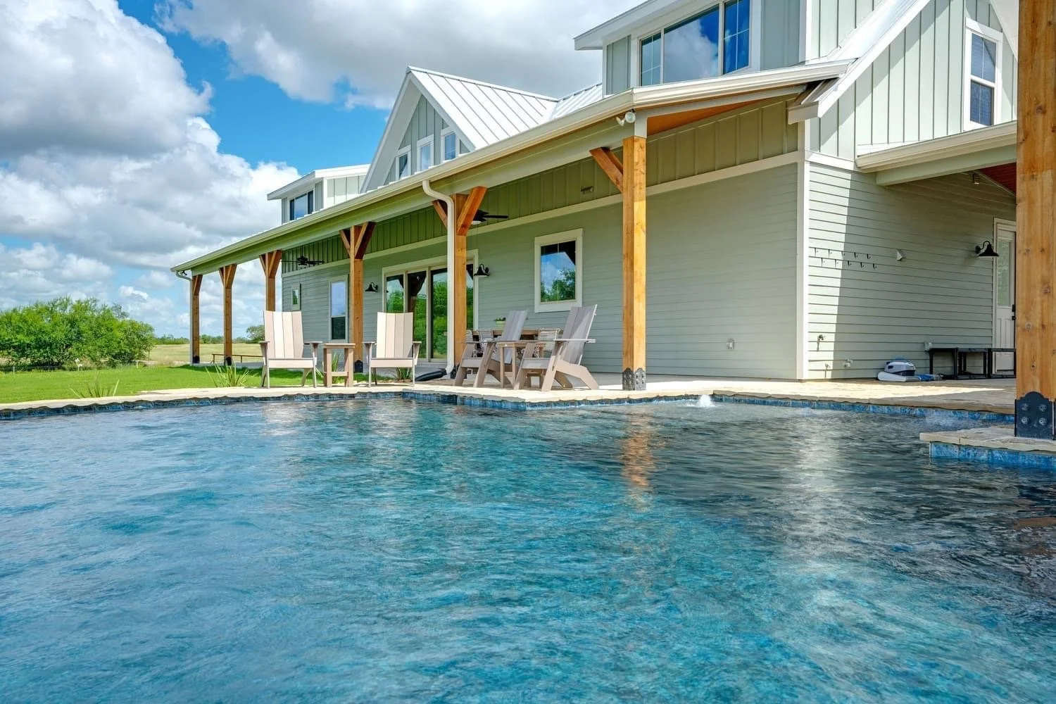 A backyard with an in-ground swimming pool next to a house with a covered patio area, outdoor seating, and a grassy lawn with trees in the background under a partly cloudy sky.