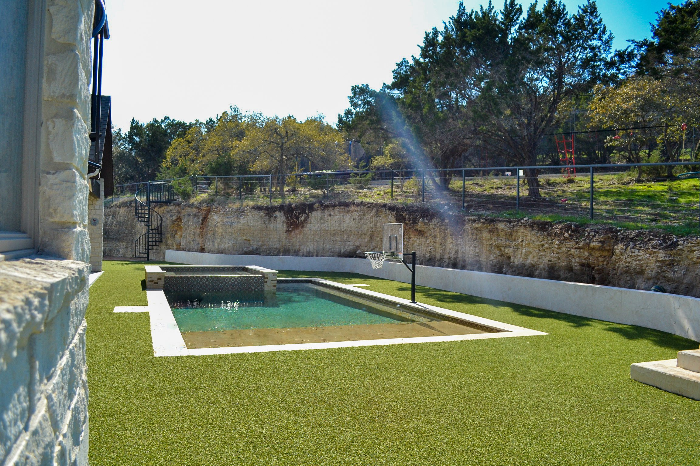 A backyard with a small swimming pool, artificial grass, and a basketball hoop, surrounded by a stone wall and trees.