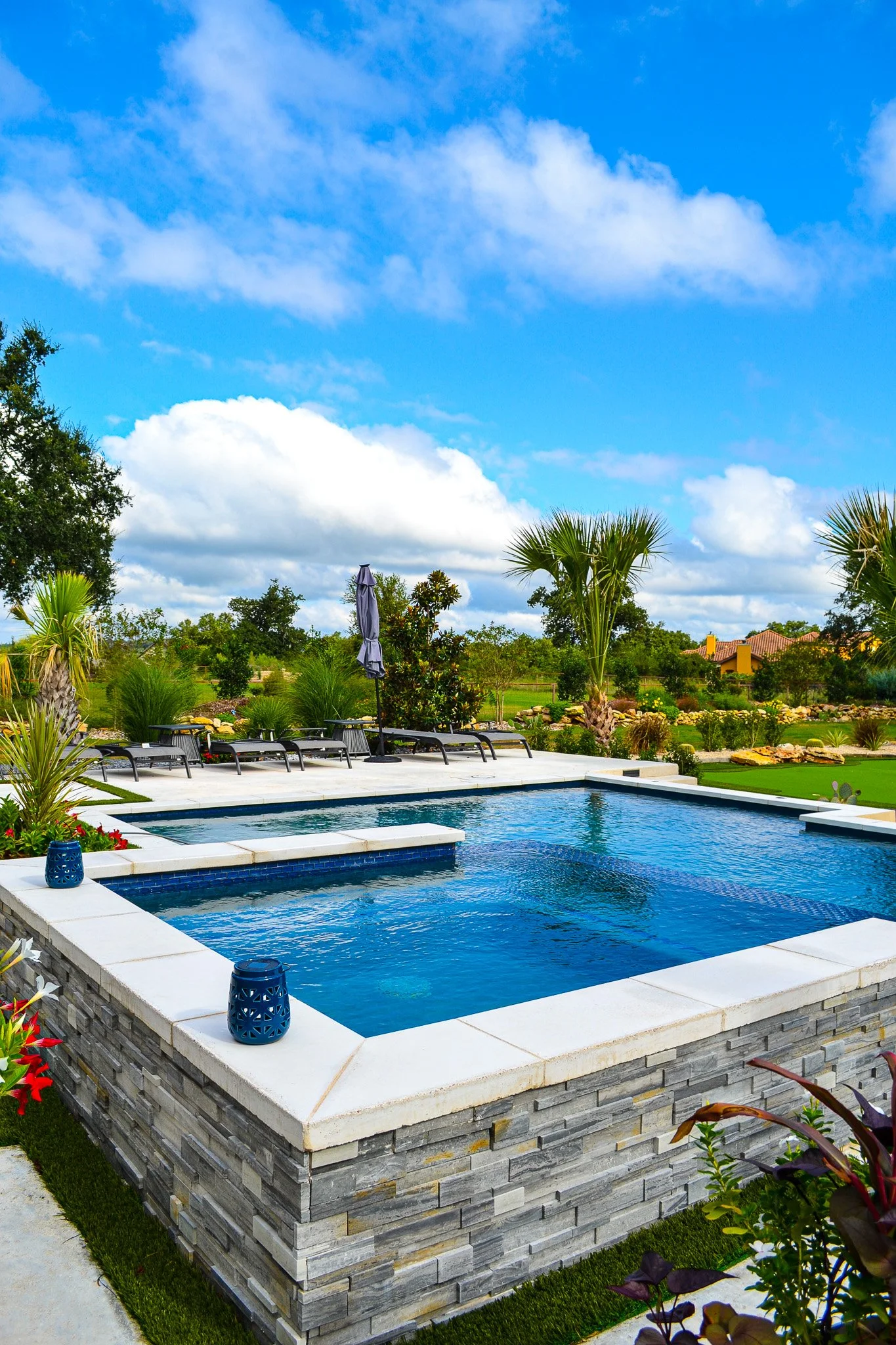 A backyard swimming pool with a built-in hot tub, surrounded by lounge chairs, lush plants, palm trees, and a blue sky with fluffy clouds.