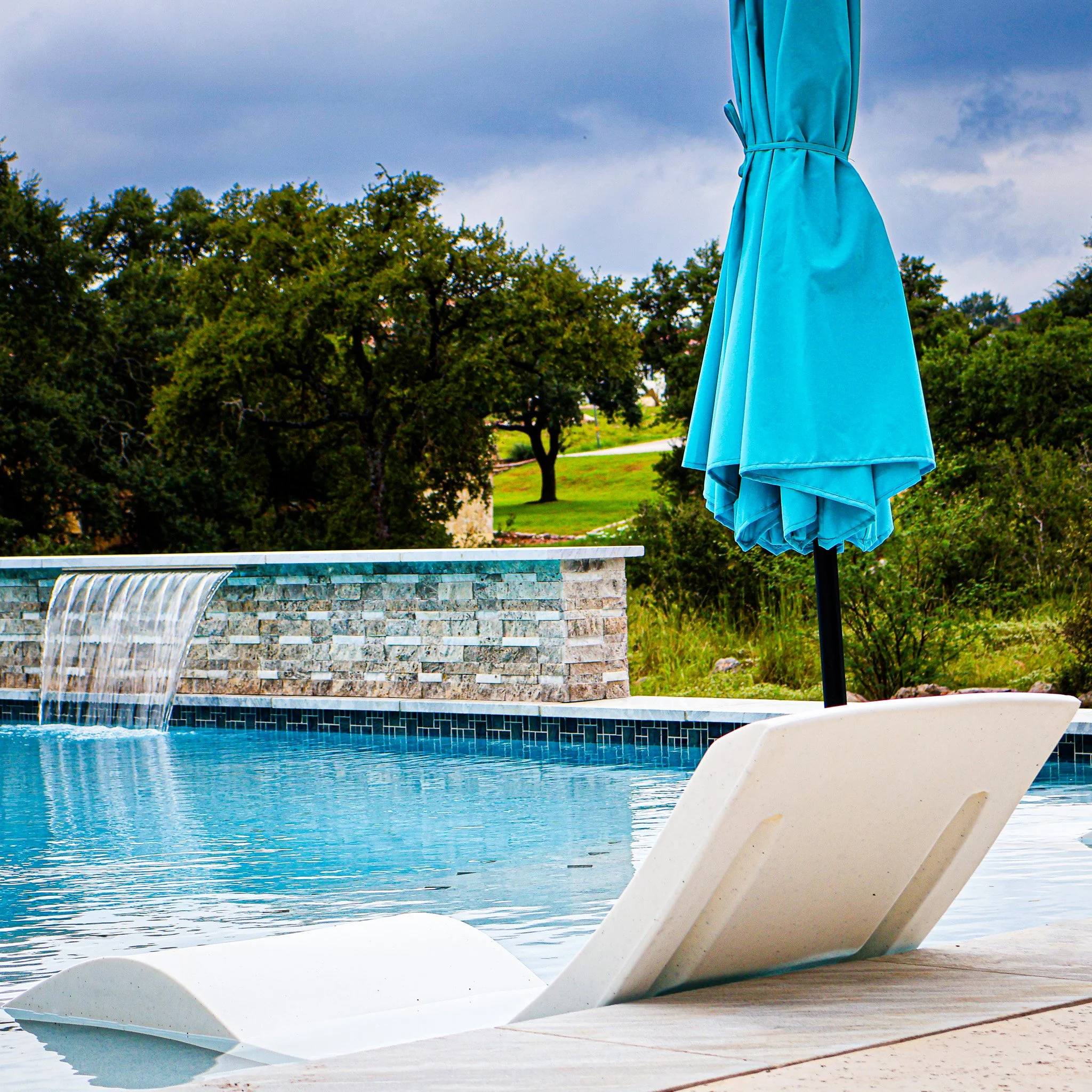 Poolside scene with a white lounge chair, closed blue umbrella, and a swimming pool with a waterfall feature, surrounded by greenery.