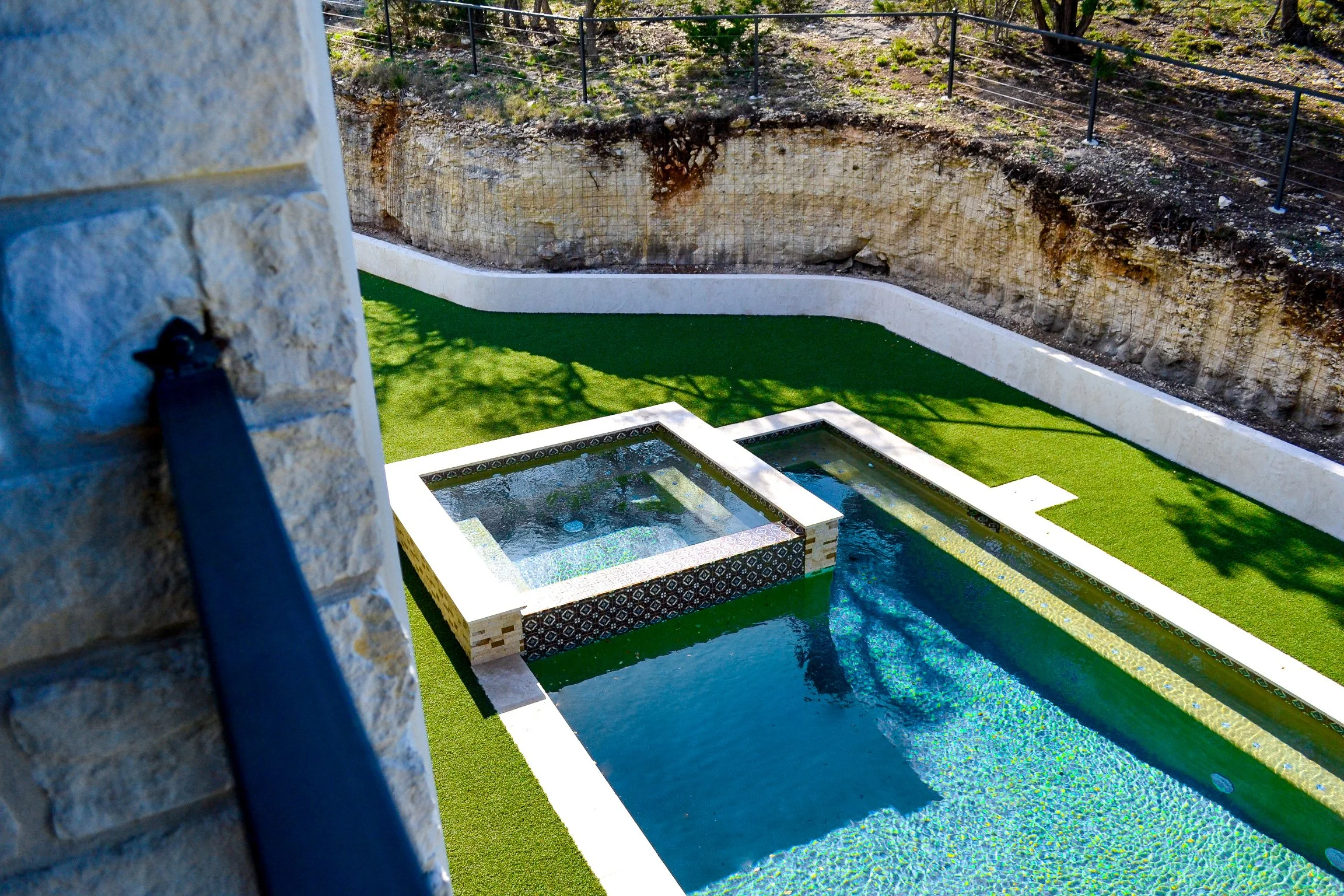 A modern backyard with a swimming pool and a hot tub, surrounded by artificial grass and a stone wall.