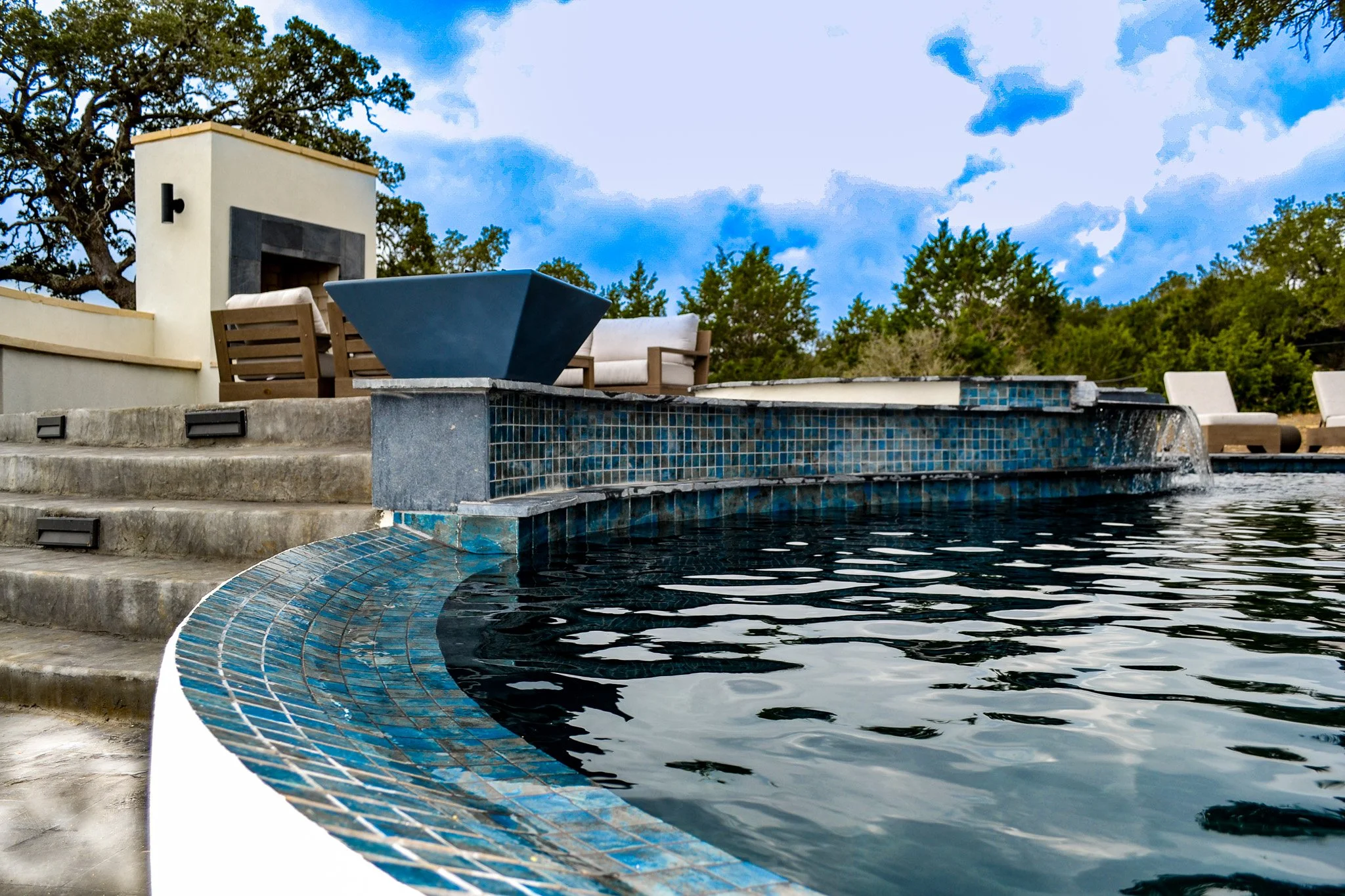 Outdoor swimming pool with blue mosaic tiles, surrounded by concrete steps and lounge chairs, with a fire pit and trees in the background under a partly cloudy sky.