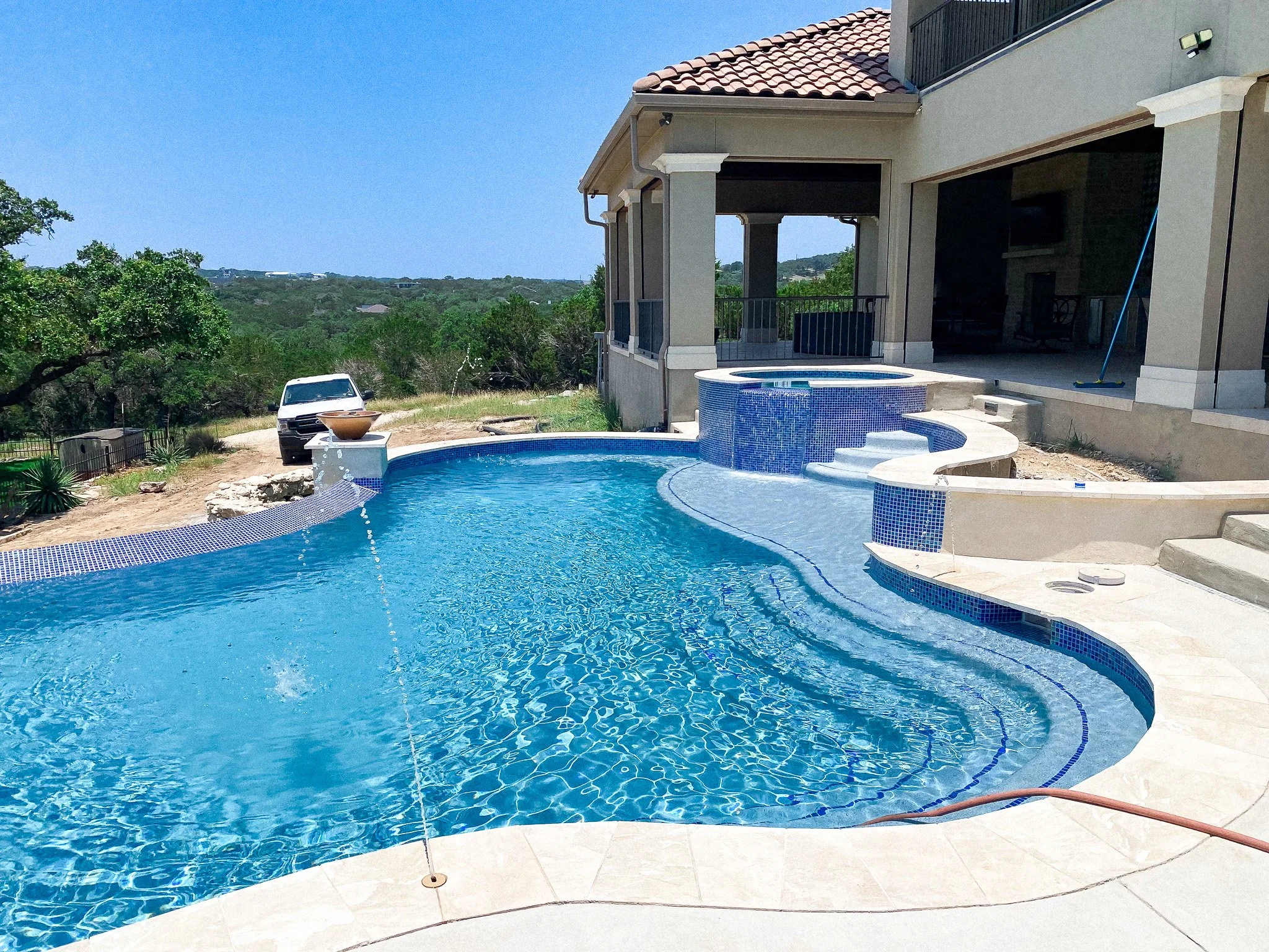 Newly constructed backyard with a freeform swimming pool, hot tub, and patio area next to a house, with a car parked on dirt and a scenic view of greenery and blue sky in the background.