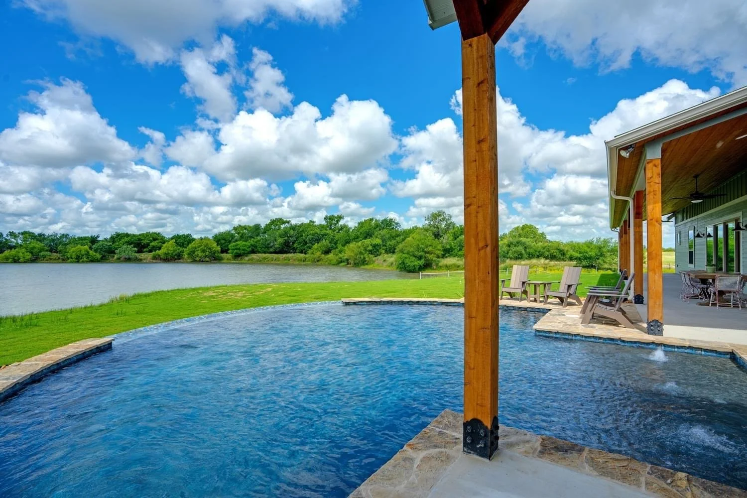 Outdoor view of a backyard with a small swimming pool, lawn, a river in the background, a blue sky with clouds, and a house with a covered patio and chairs.