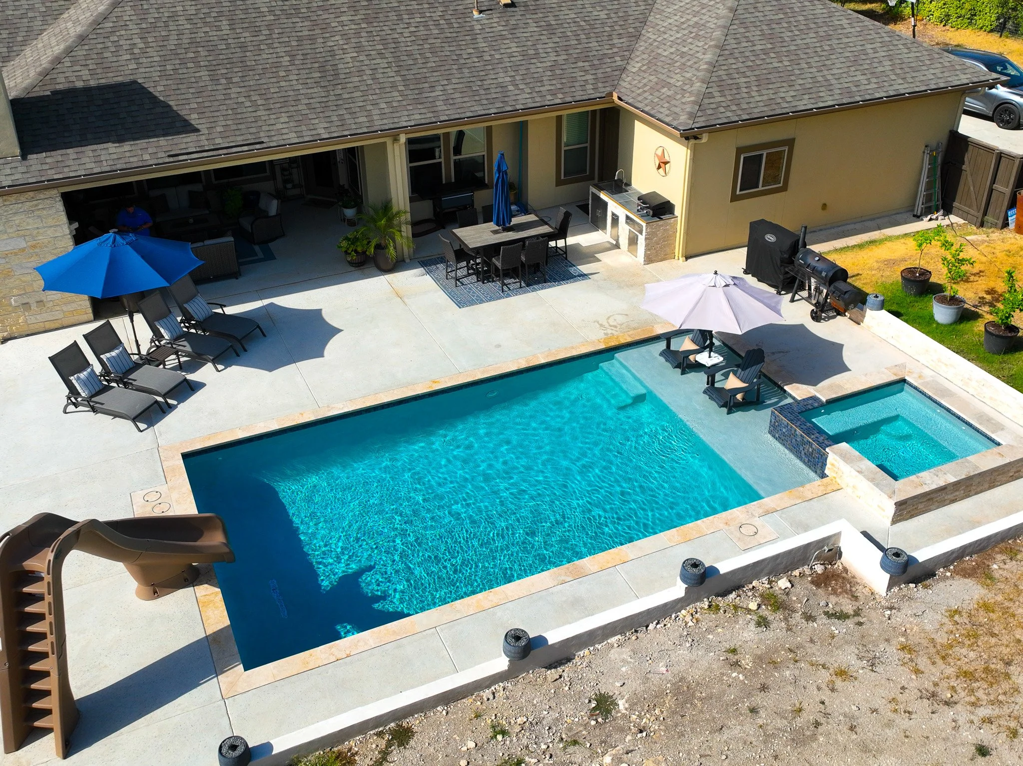 A backyard with a swimming pool, a hot tub, lounge chairs with umbrellas, a slide, and an outdoor kitchen area with a dining table and chairs.