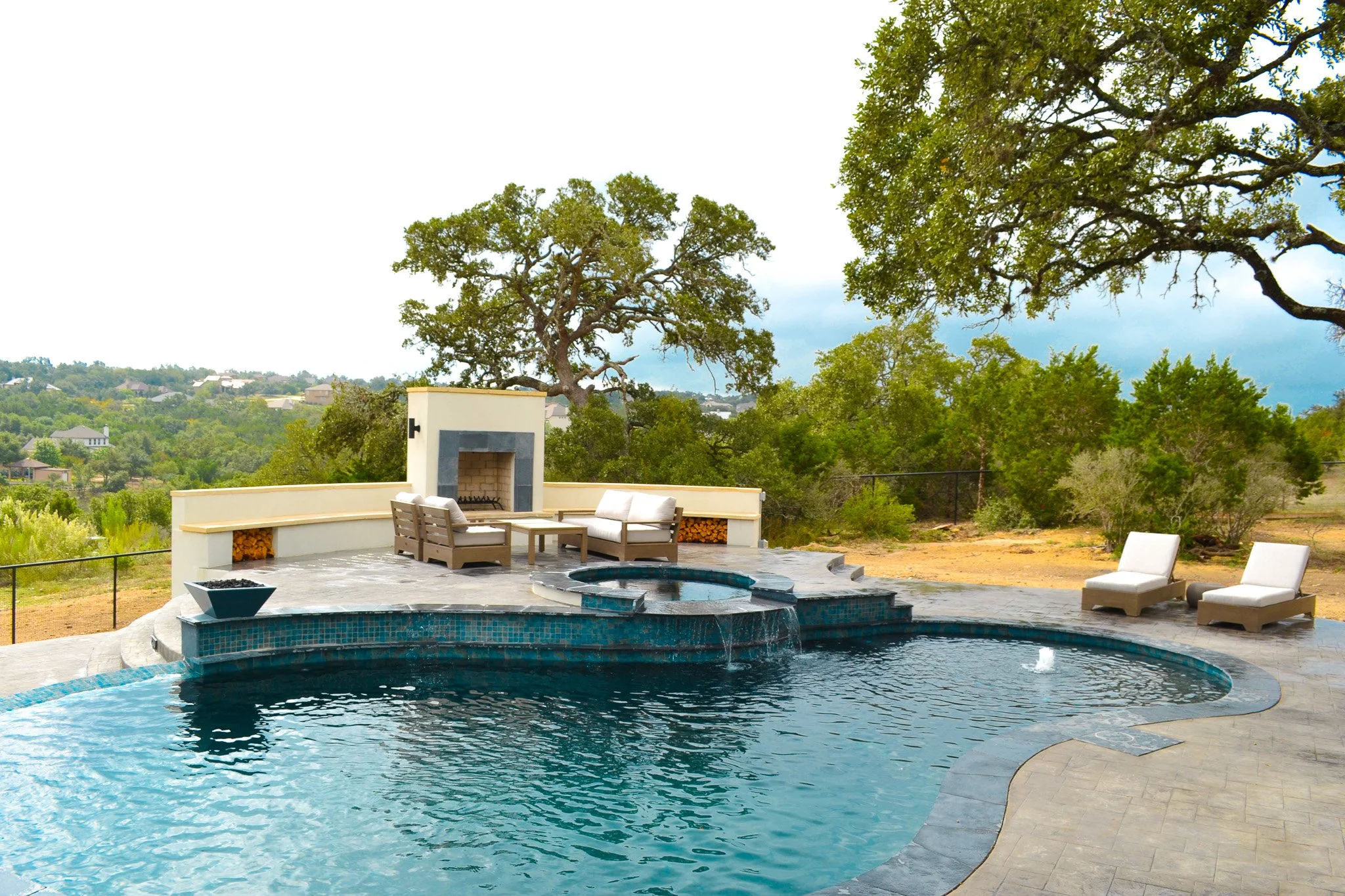 A backyard swimming pool with a hot tub, poolside chairs, and a fireplace under trees with a scenic view.